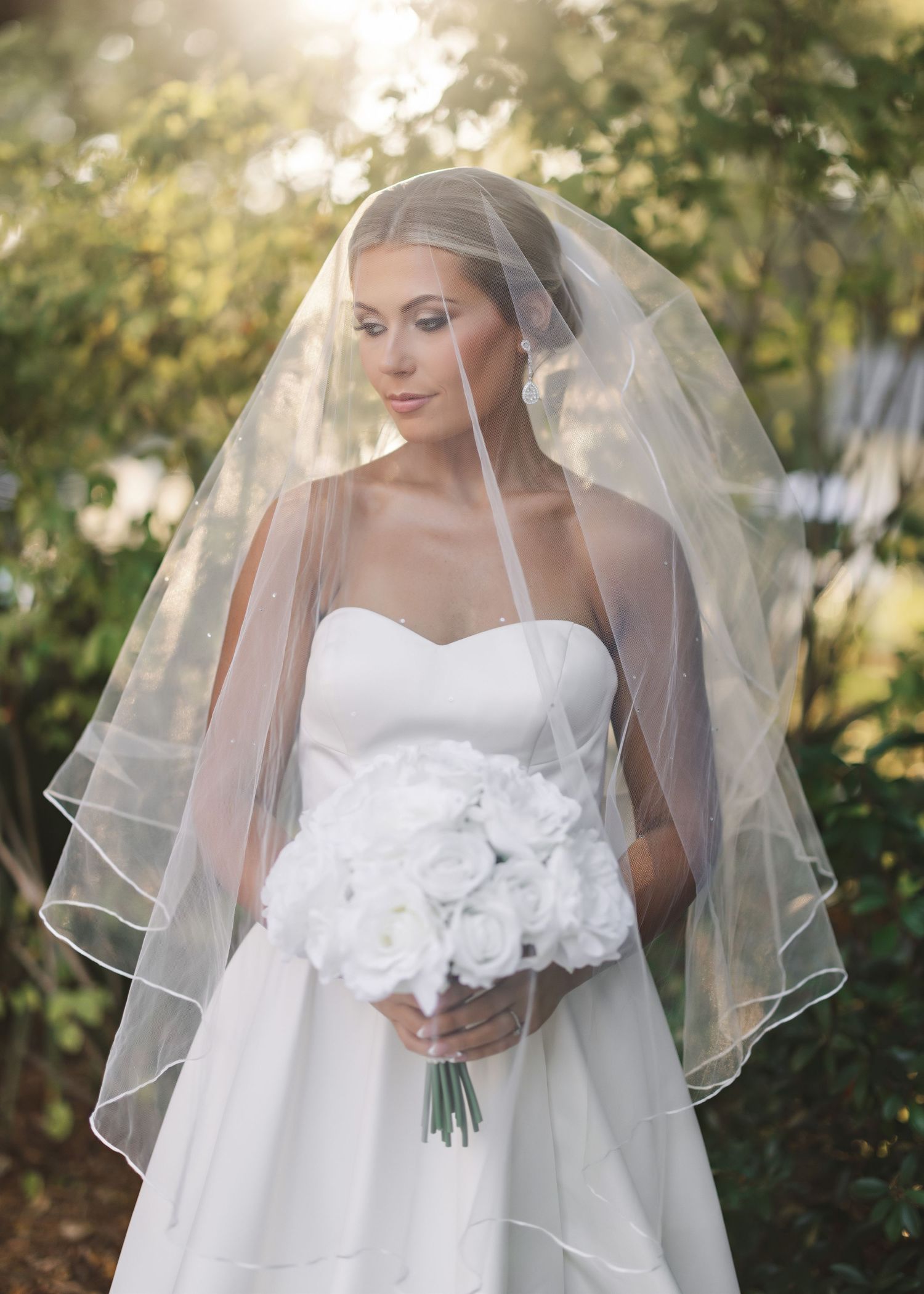 A bride in a white wedding dress and flowing veil holds a bouquet in an outdoor garden setting.