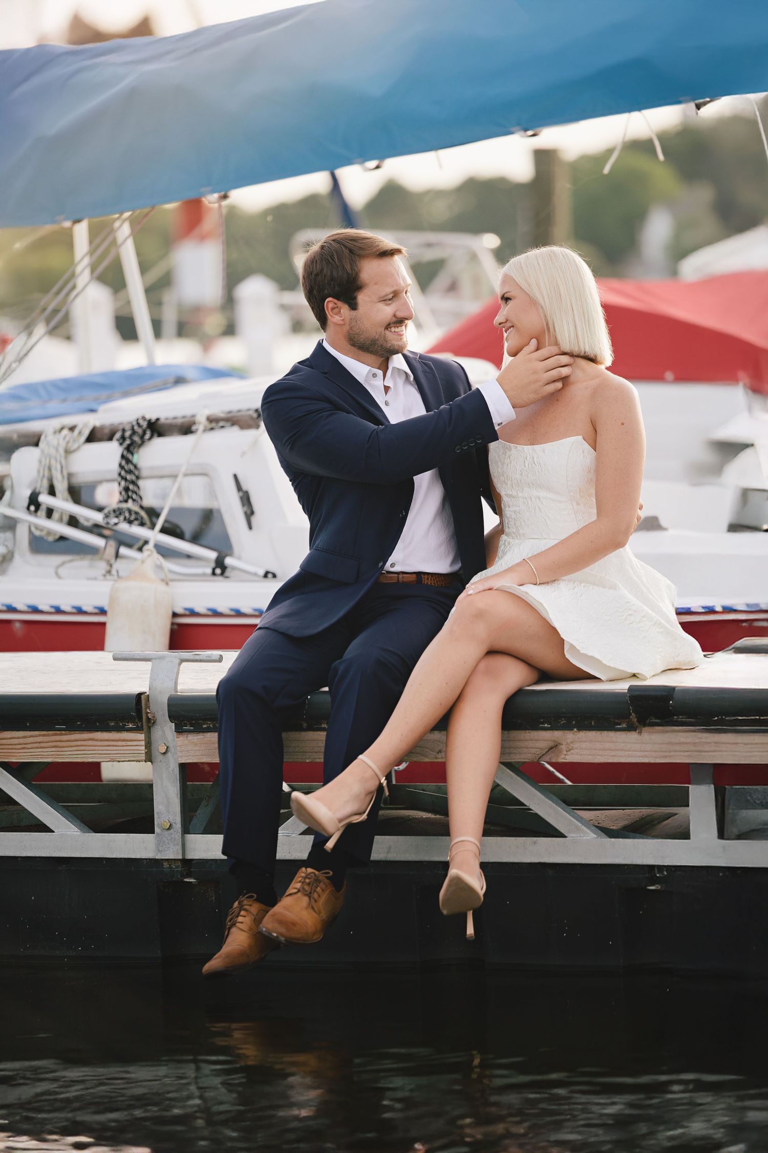 Romantic couple sitting on dock at marina with boats and water in background during sunset engagement photoshoot.