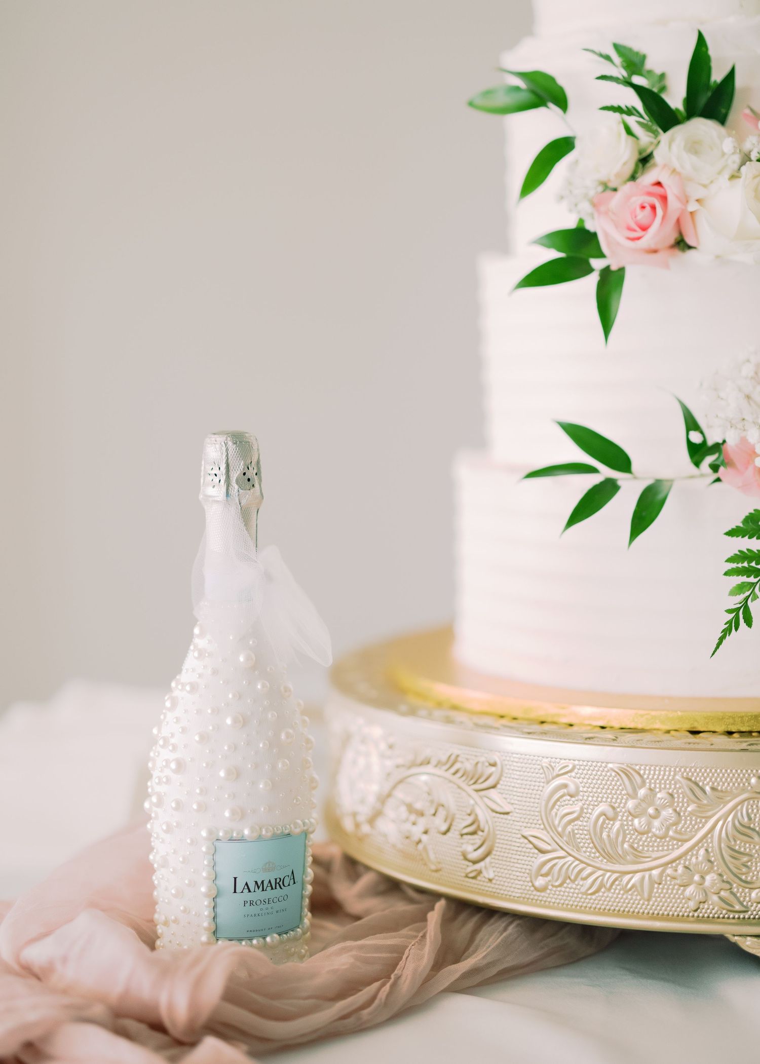 Elegant white wedding cake decorated with pink roses and green foliage sits on ornate gold stand.