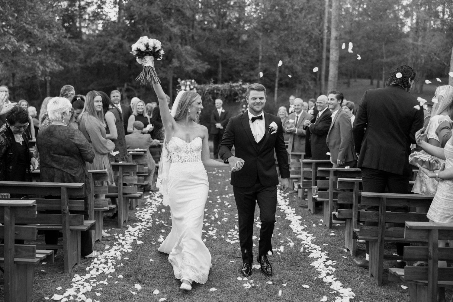 Two newlyweds celebrate joyfully while walking down an outdoor aisle lined with rose petals at a garden wedding ceremony.