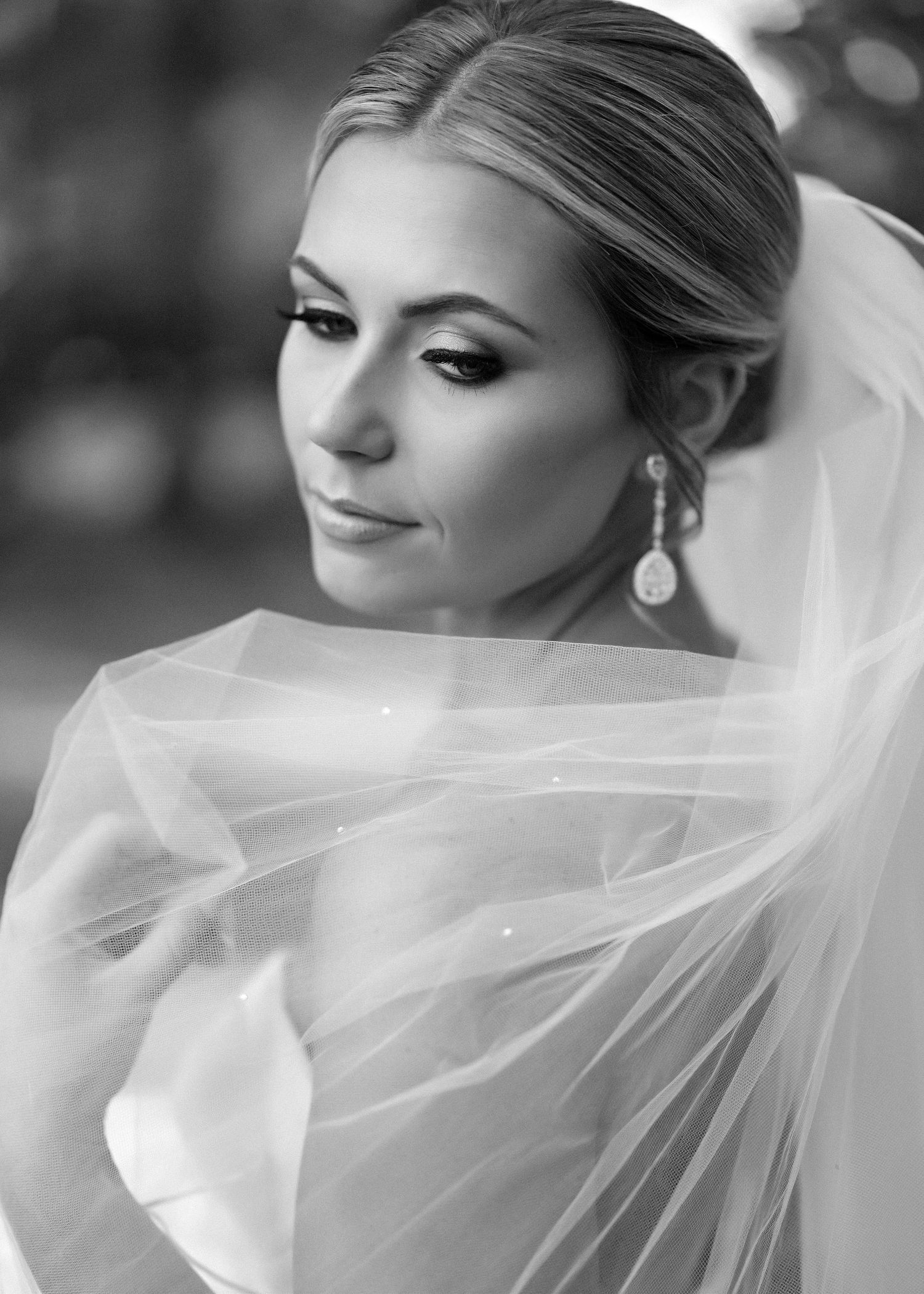 A black and white portrait of a bride wearing a veil and dangling earrings, looking to the side with elegant makeup.