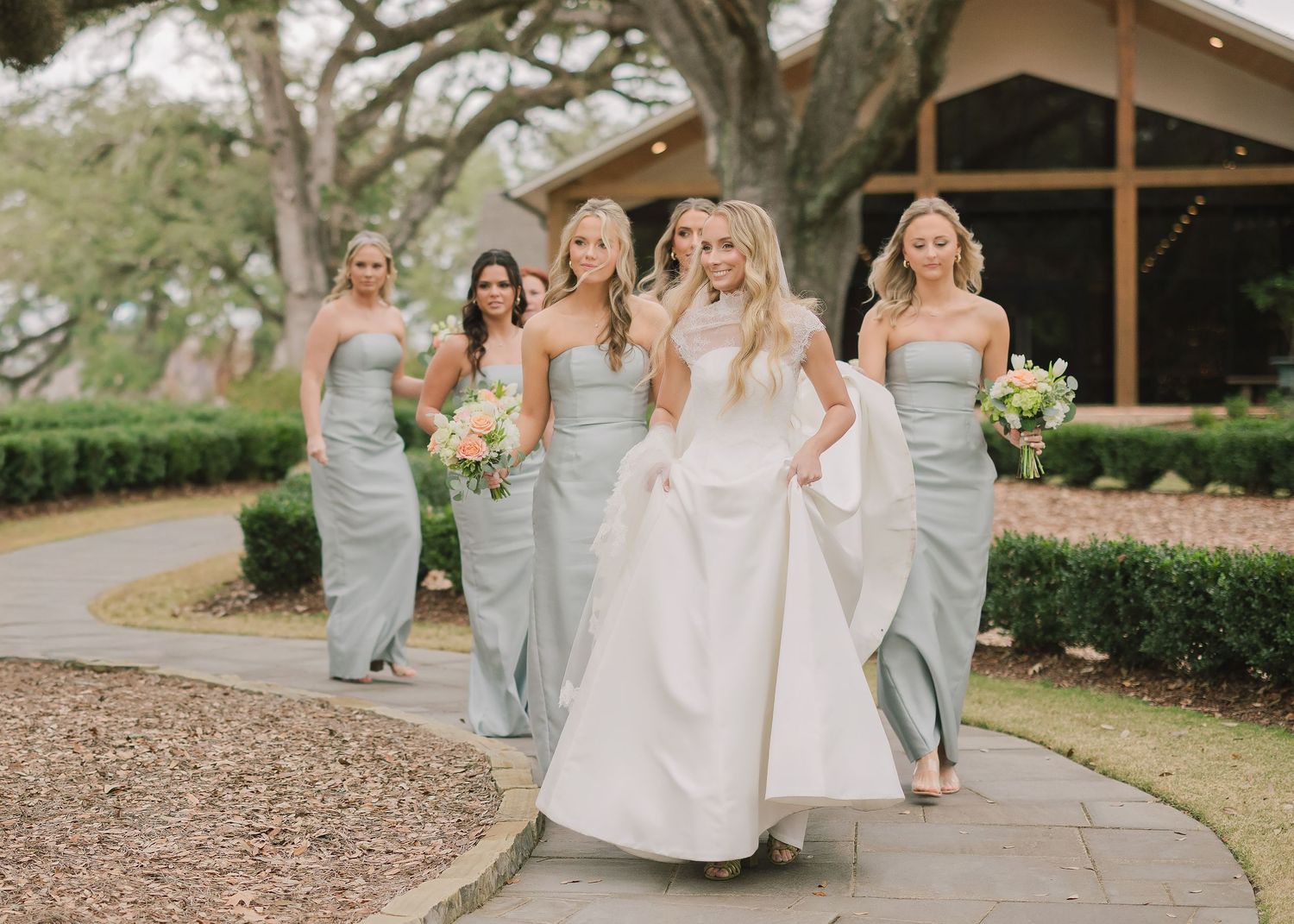 A bride in a white gown walks with bridesmaids in silver dresses along a curved path outside a rustic venue.