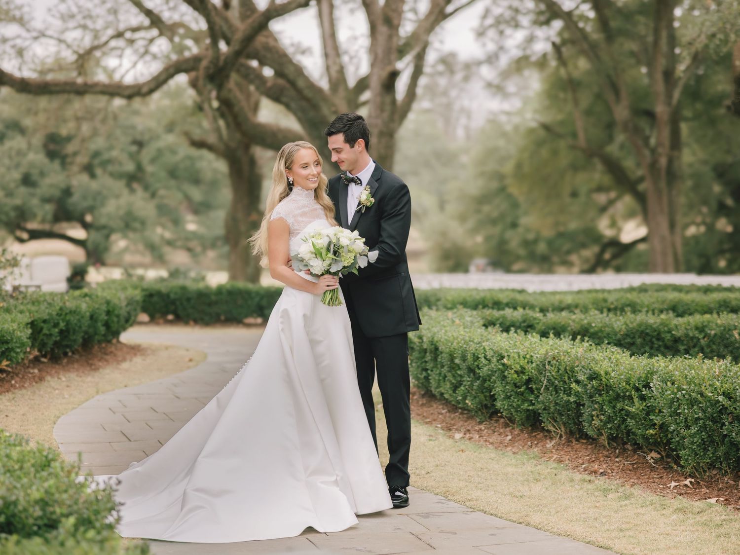 A couple in wedding attire poses romantically along a tree-lined pathway in a garden setting.