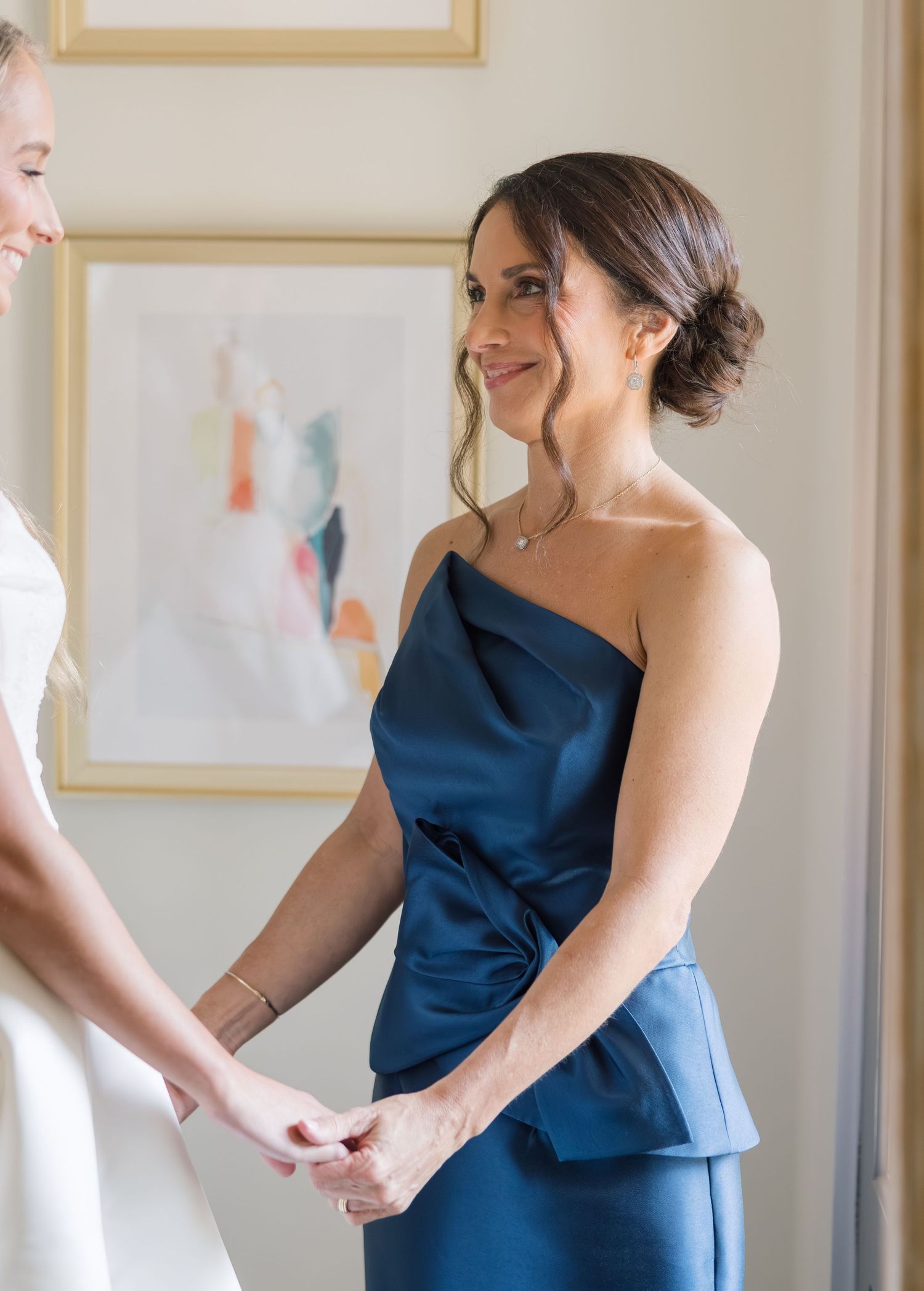 A person in a navy blue formal dress smiles and gestures while standing in an elegant room with framed artwork.