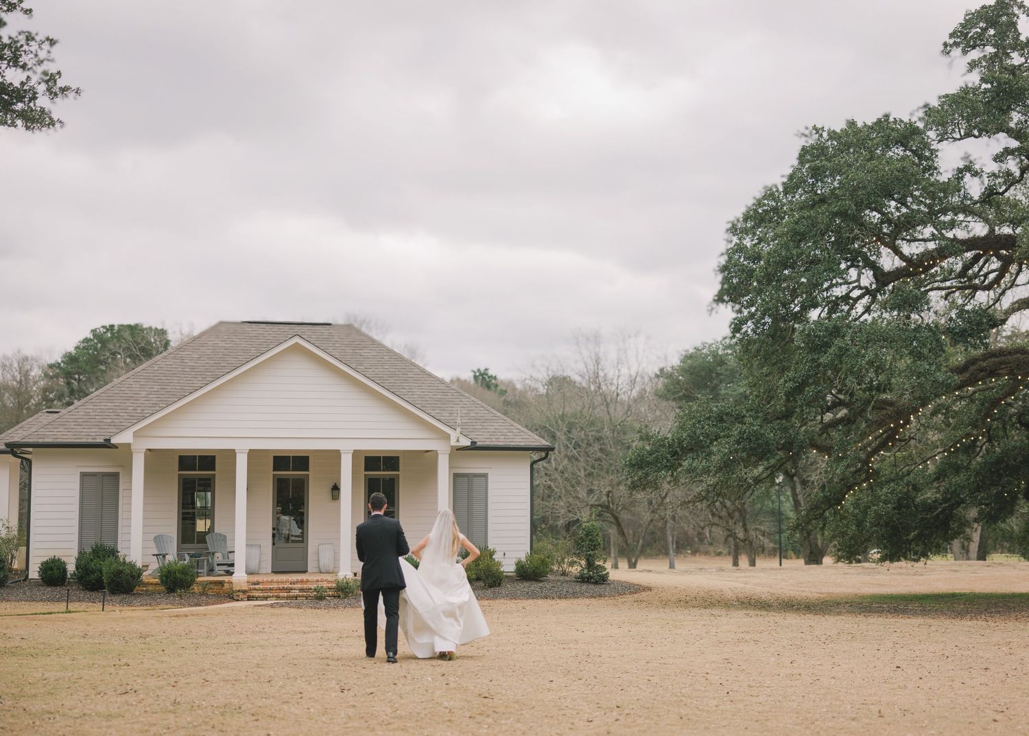 A panoramic view of a rustic wedding ceremony setup with white chairs on a grassy field surrounded by oak trees.