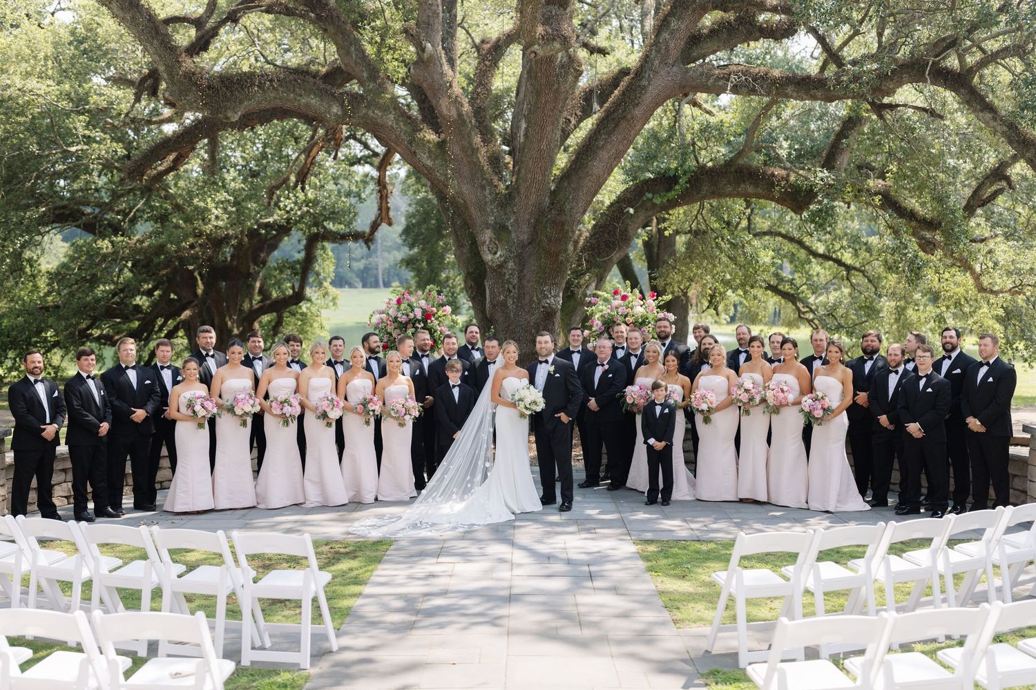 Outdoor wedding ceremony setup beneath majestic oak trees with white chairs and floral arrangements.