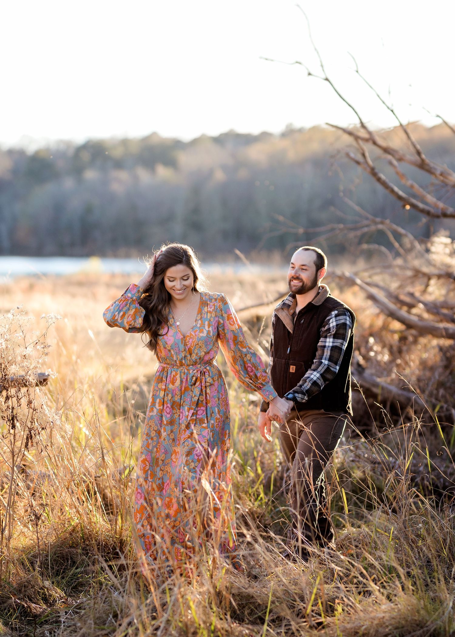 Couple walking through golden tall grass near a lake during sunset.