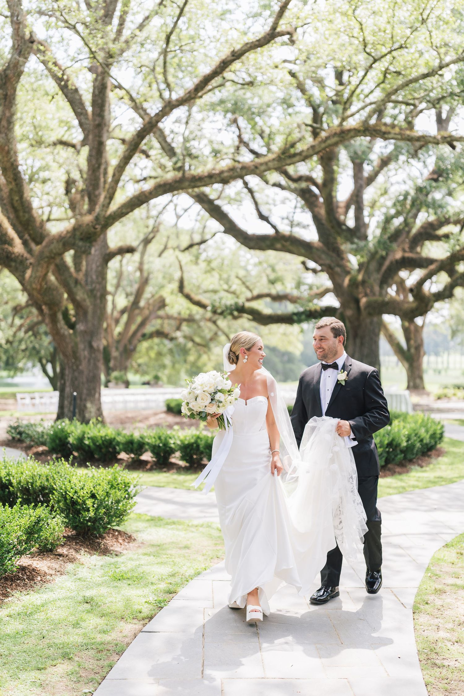 Elegant couple strolls beneath oak trees on wedding day path.