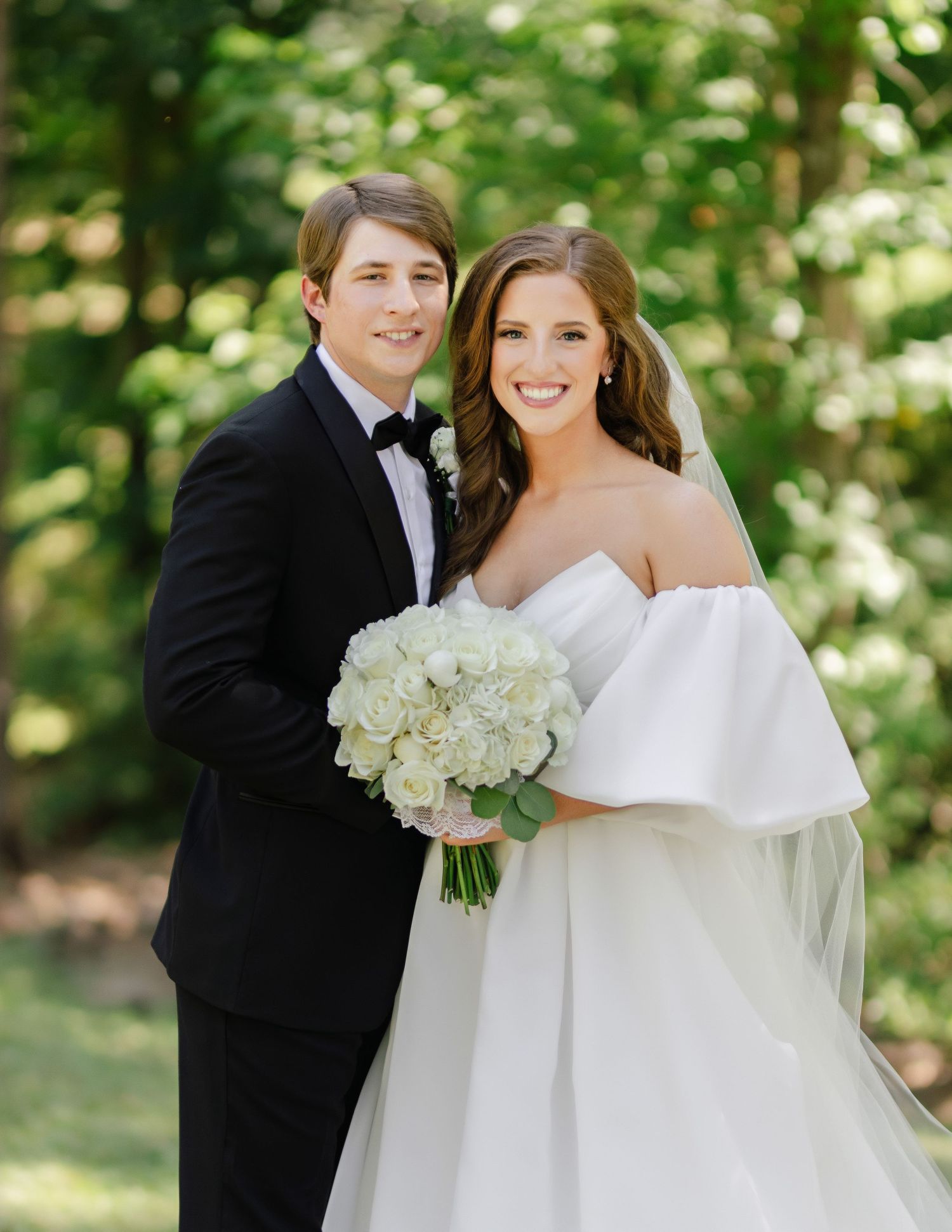 Newlyweds pose in formal attire with white bouquet in garden setting.