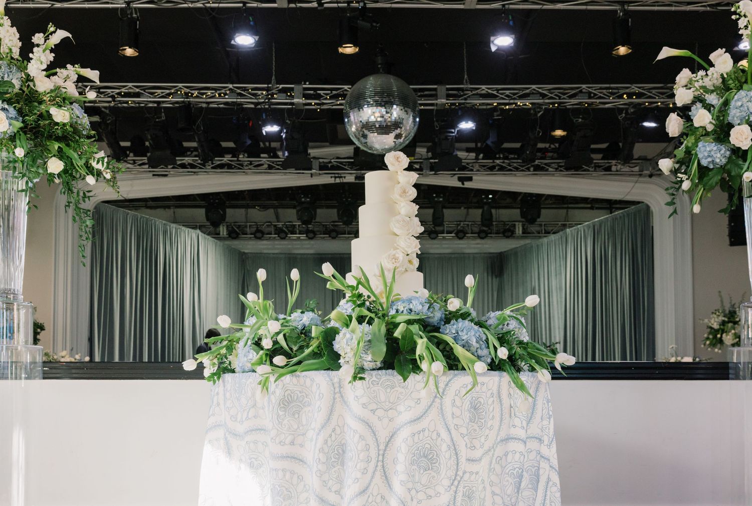 Elegant white wedding cake display with floral arrangements and green accents on draped table.