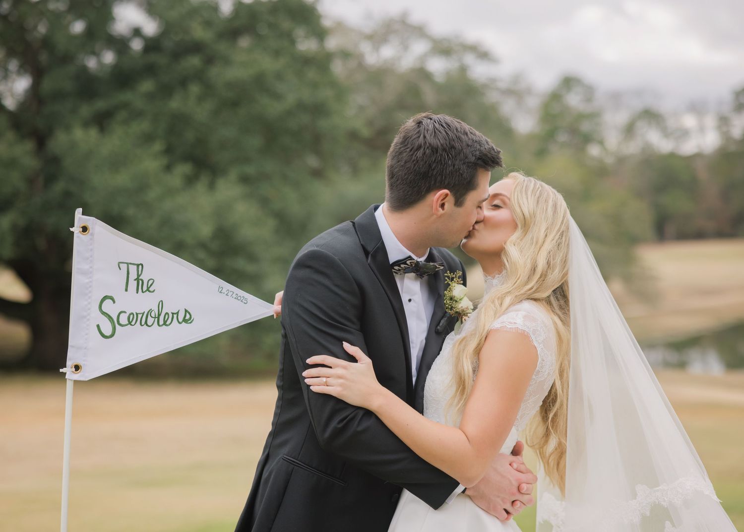 A romantic wedding couple shares intimate kisses in an outdoor garden setting with a flag marker in the background.