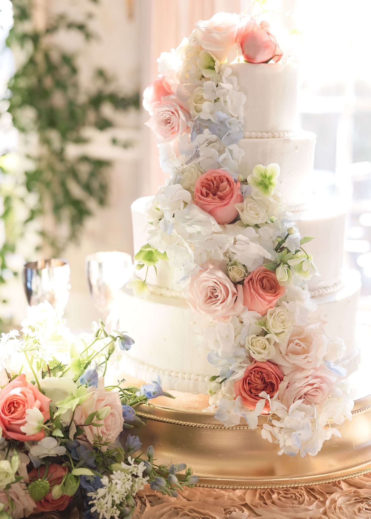Wedding cake decorated with blush pink roses and white hydrangeas on golden stand.