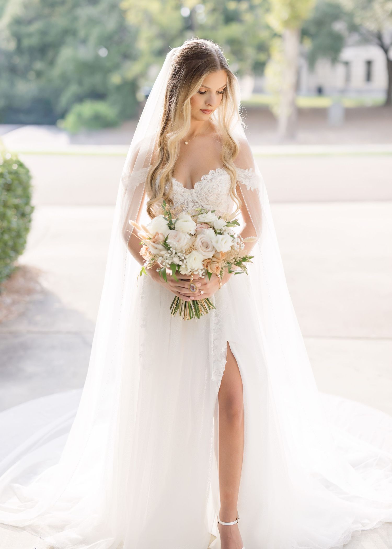 A bride in a flowing white dress holds a romantic bouquet of white roses and greenery in a sun-filled outdoor setting.