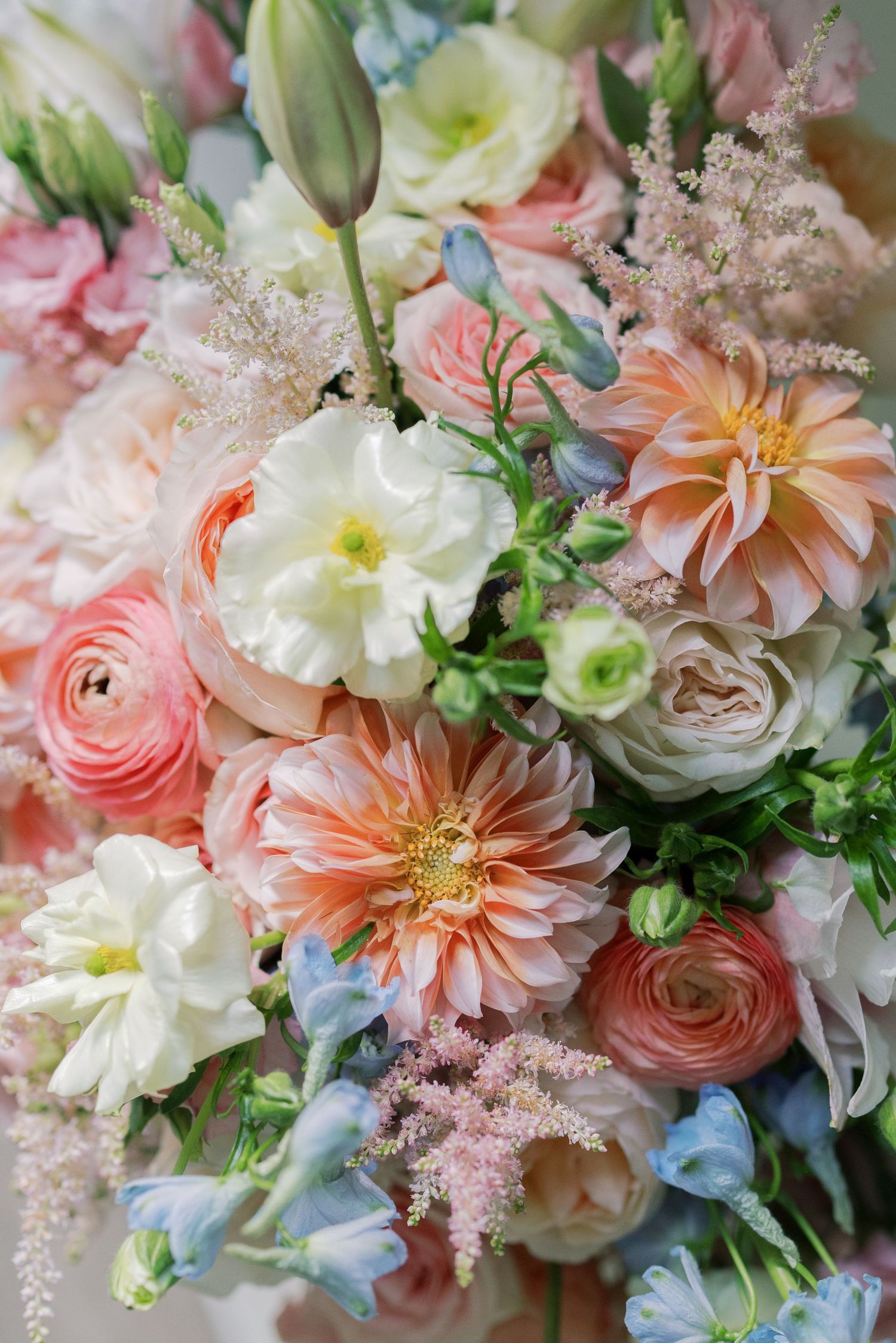 A romantic floral arrangement featuring dahlias, ranunculus, roses and delphinium in soft pink, peach and white tones.