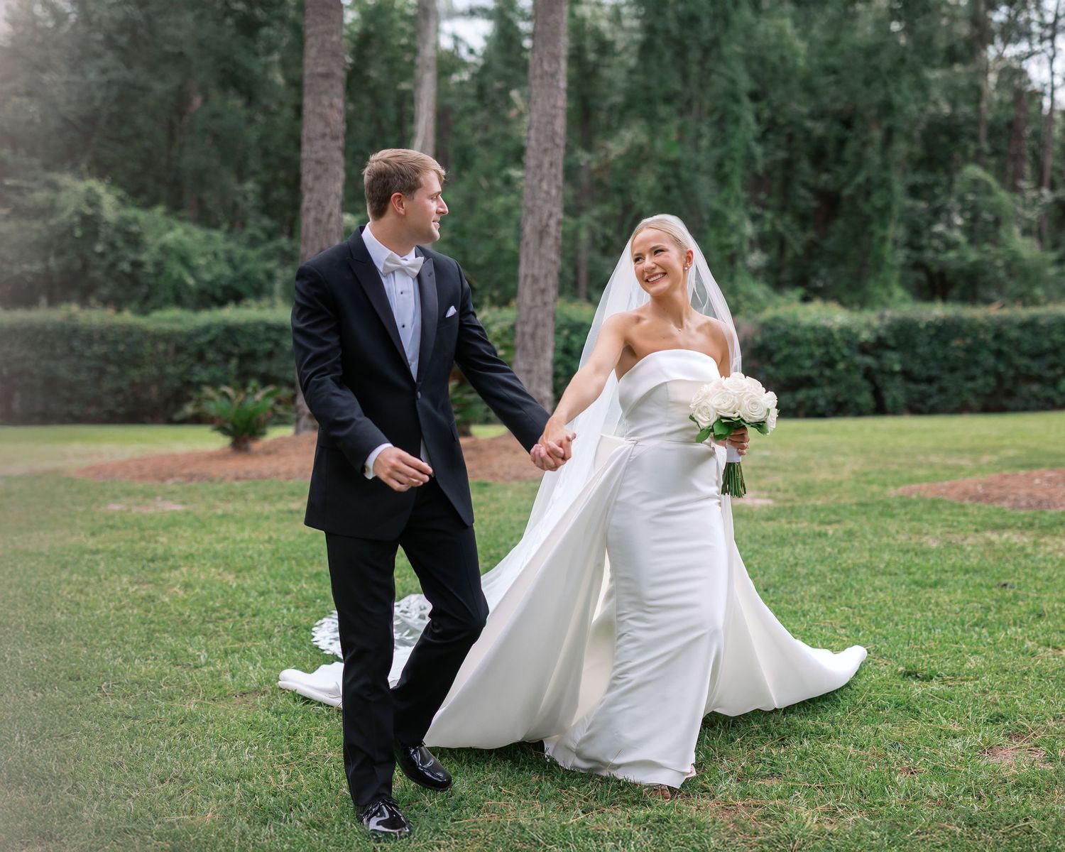 A couple in wedding attire sits together on grass in a scenic outdoor garden setting.