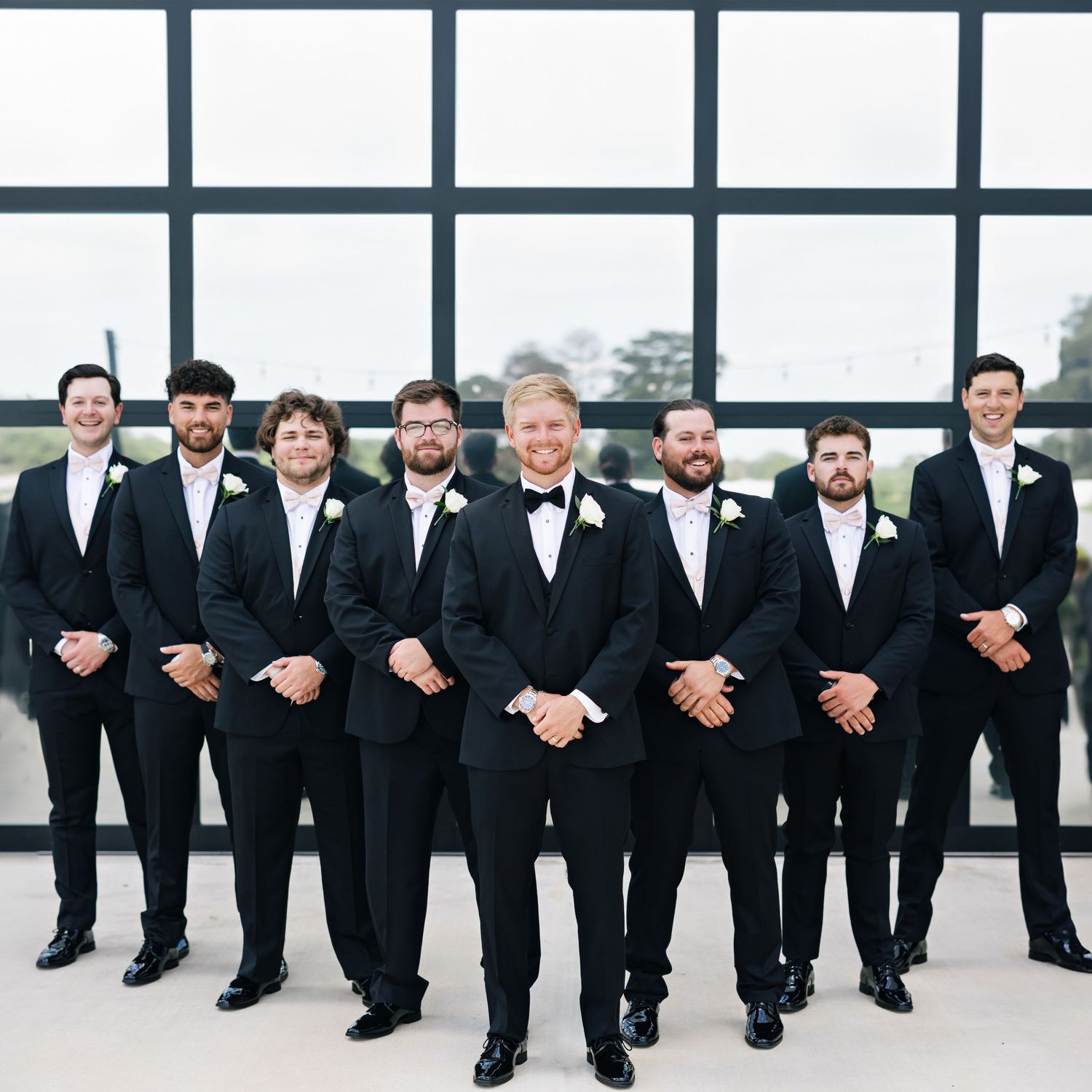 Groomsmen in black tuxedos and bowties pose together in front of modern window wall.