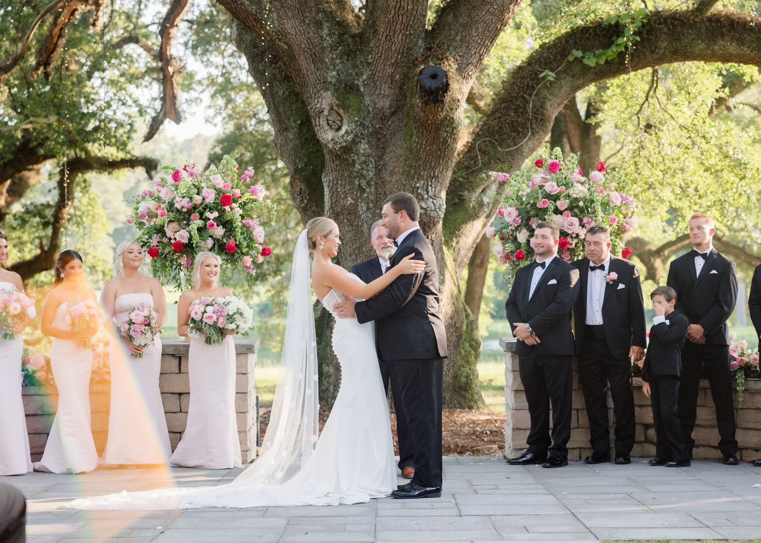 Elegant outdoor wedding ceremony under ancient oak trees with white chairs and decorative pedestals.