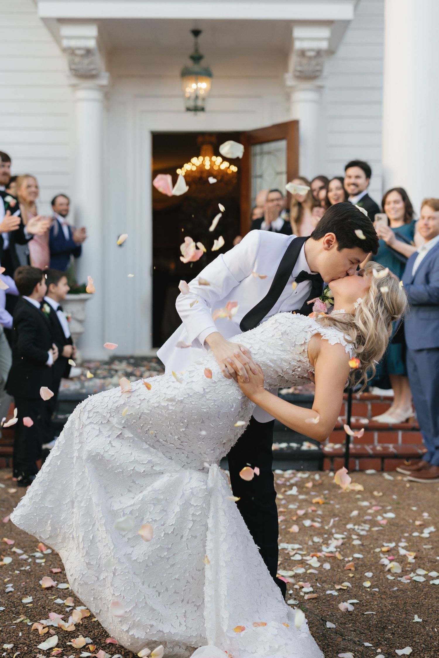 A romantic dip kiss moment outside a white columned entrance with flower petals falling.