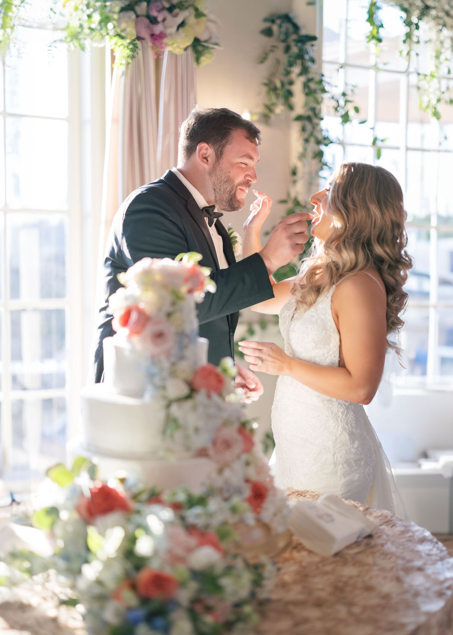 A couple shares cake and laughter during their wedding reception beside a floral decorated cake.