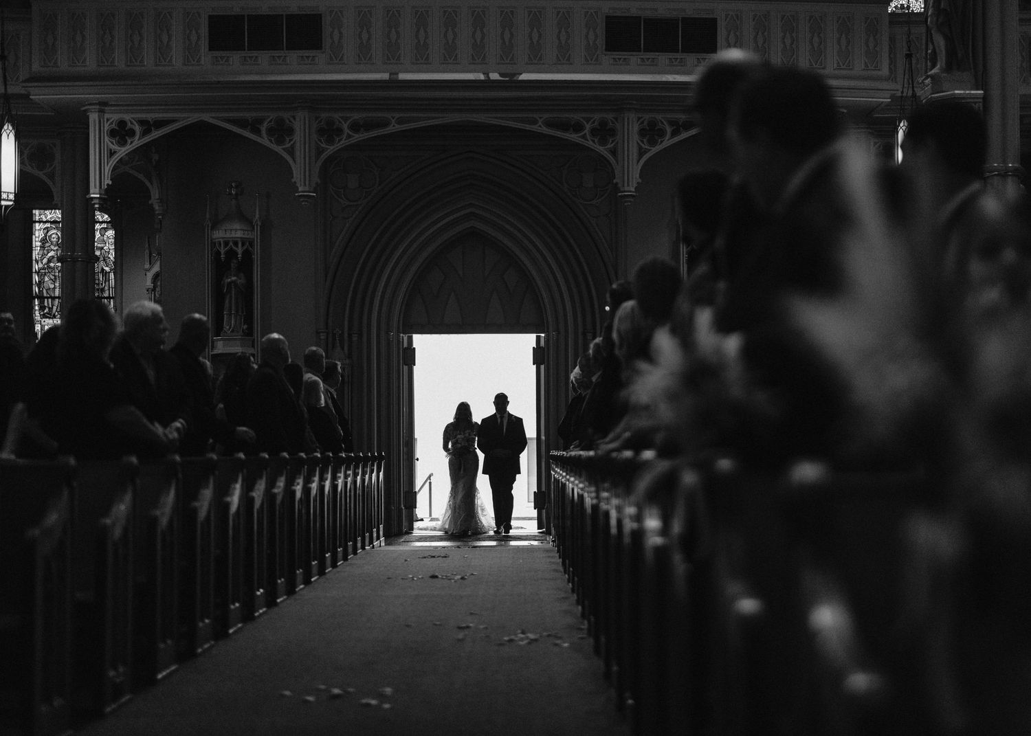 Silhouetted figures walking down church aisle in moody black and white photo.
