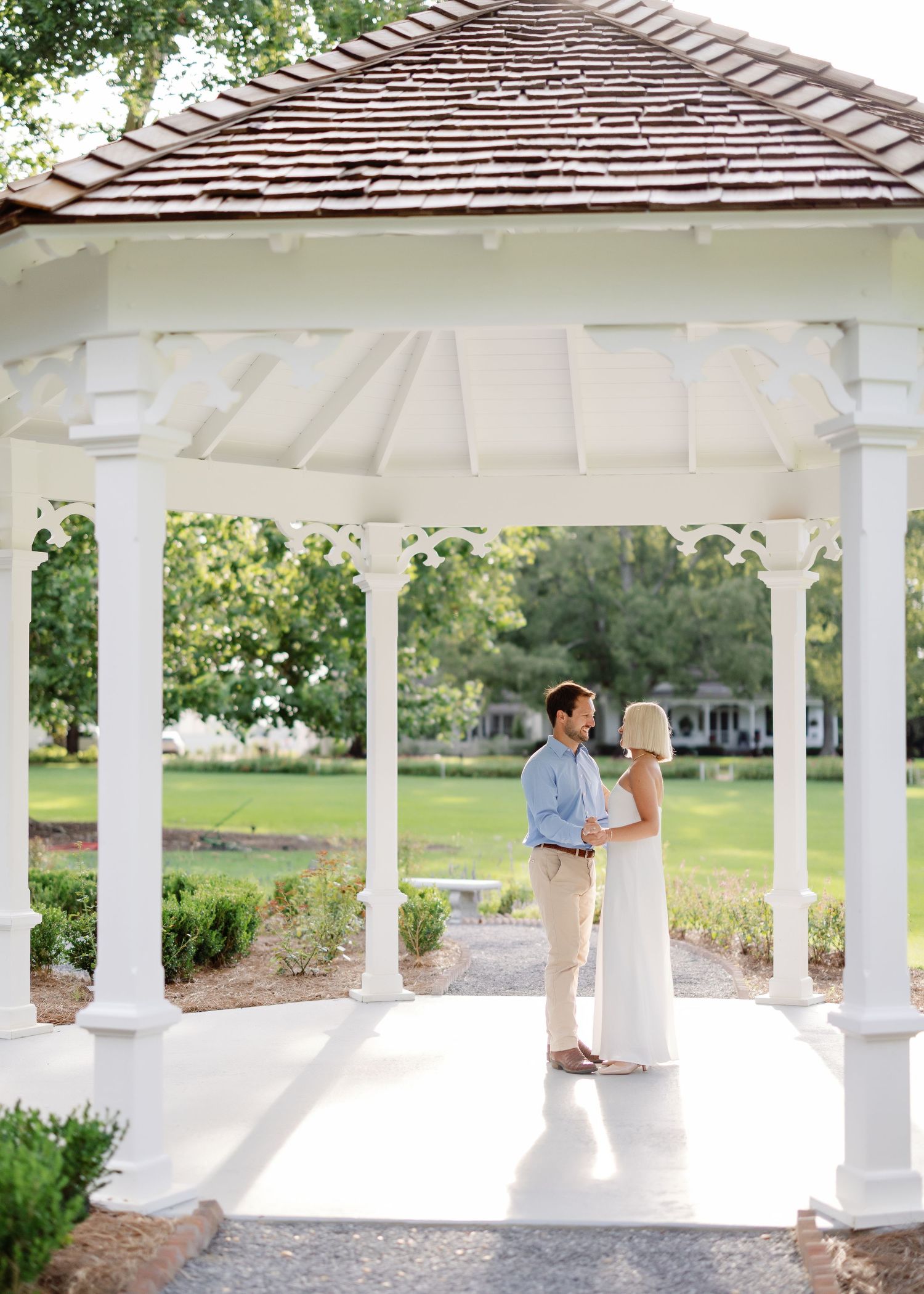 White gazebo with shingled roof in landscaped garden setting.