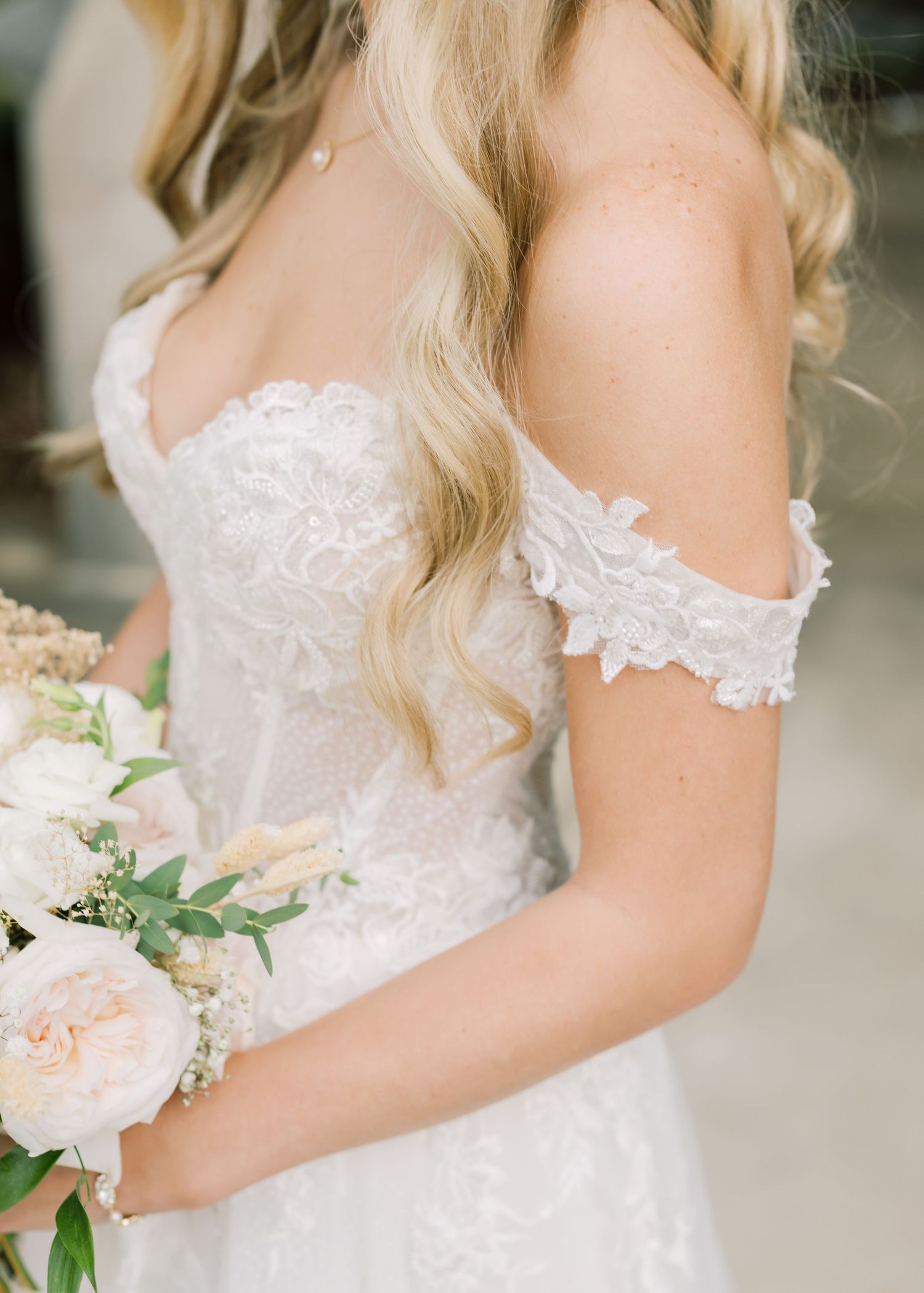 Close-up details of a bride wearing an off-shoulder lace wedding dress holding a soft pink and peach floral bouquet.