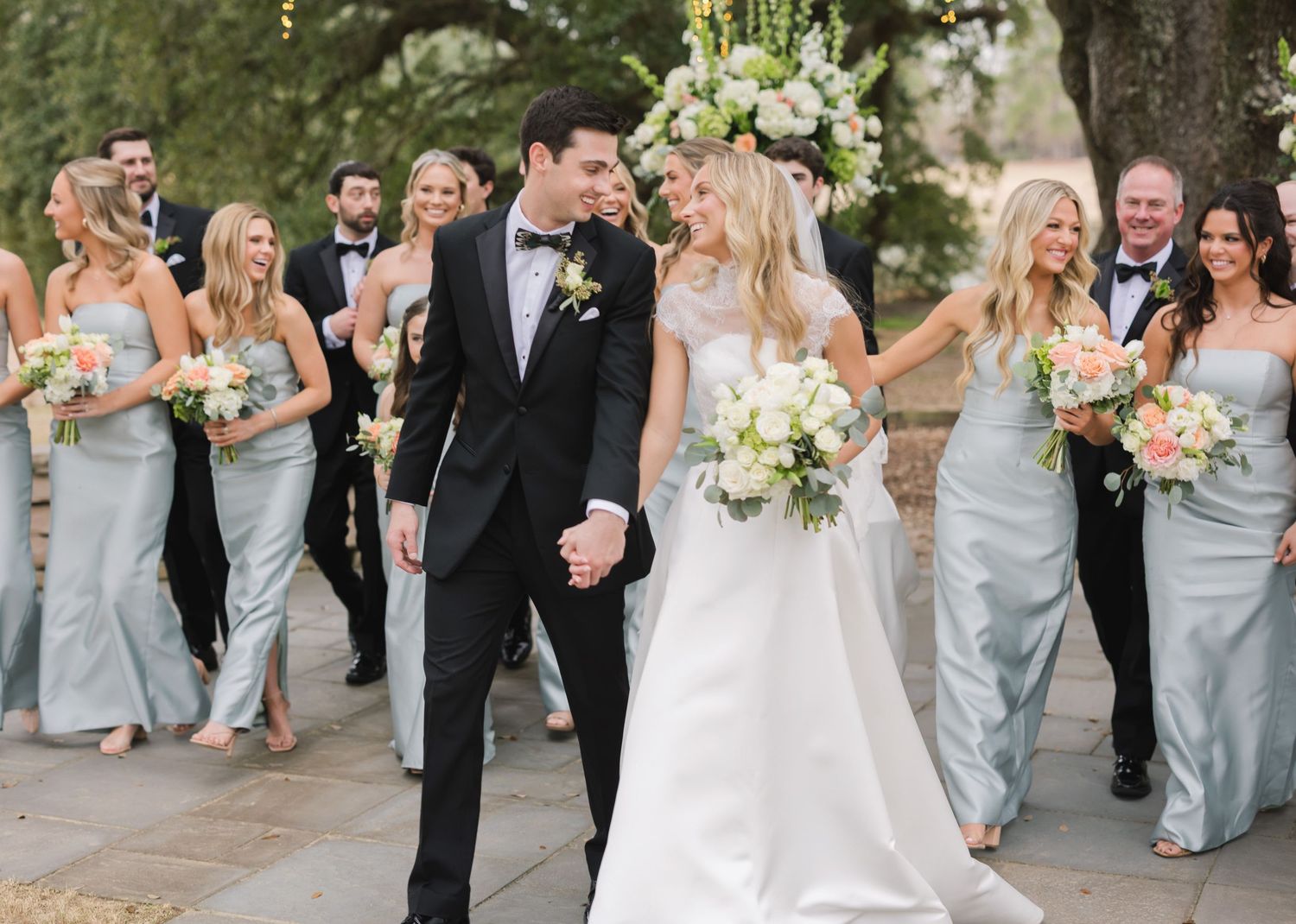 Wedding party wearing elegant gray bridesmaid dresses and black tuxedos walk together outdoors during celebration.