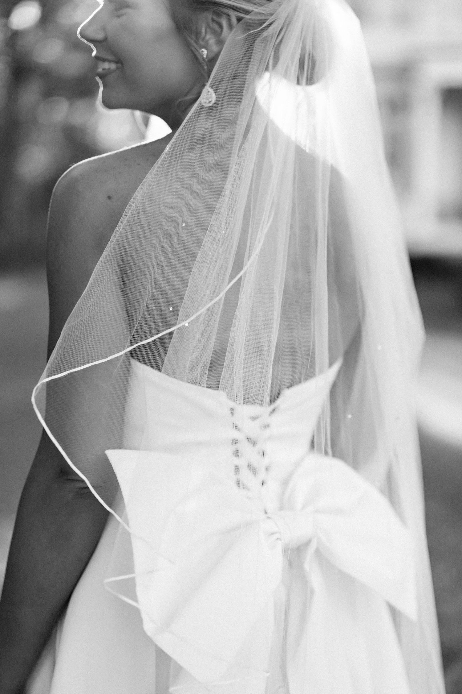 Black and white wedding photo showing elegant bridal gown details with flowing veil and large decorative bow.