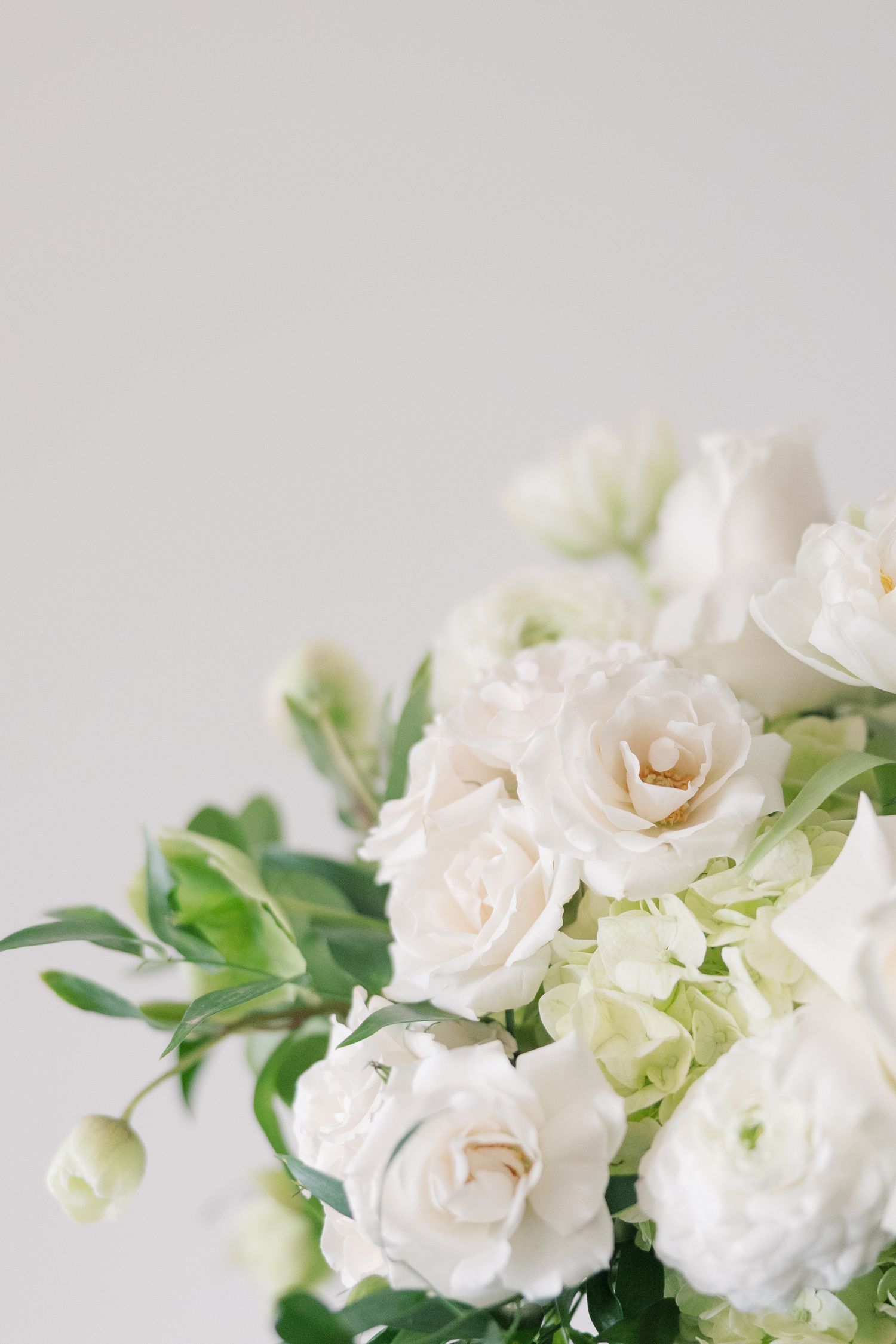 Elegant white floral bouquet featuring roses, ranunculus, and hydrangeas with lush green foliage on a soft gray background.