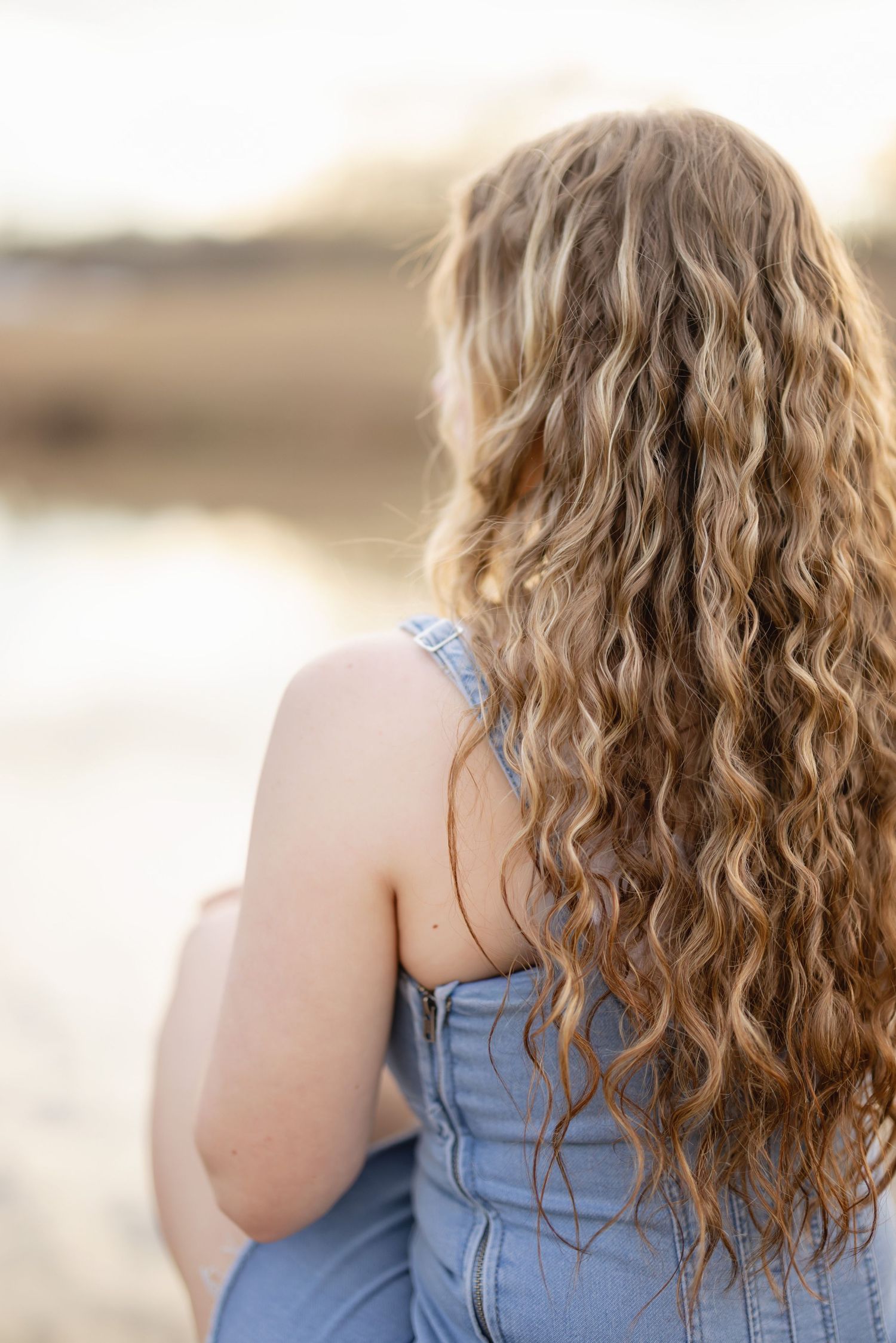 Back view of curly blonde hair and blue denim top against blurred beige background.