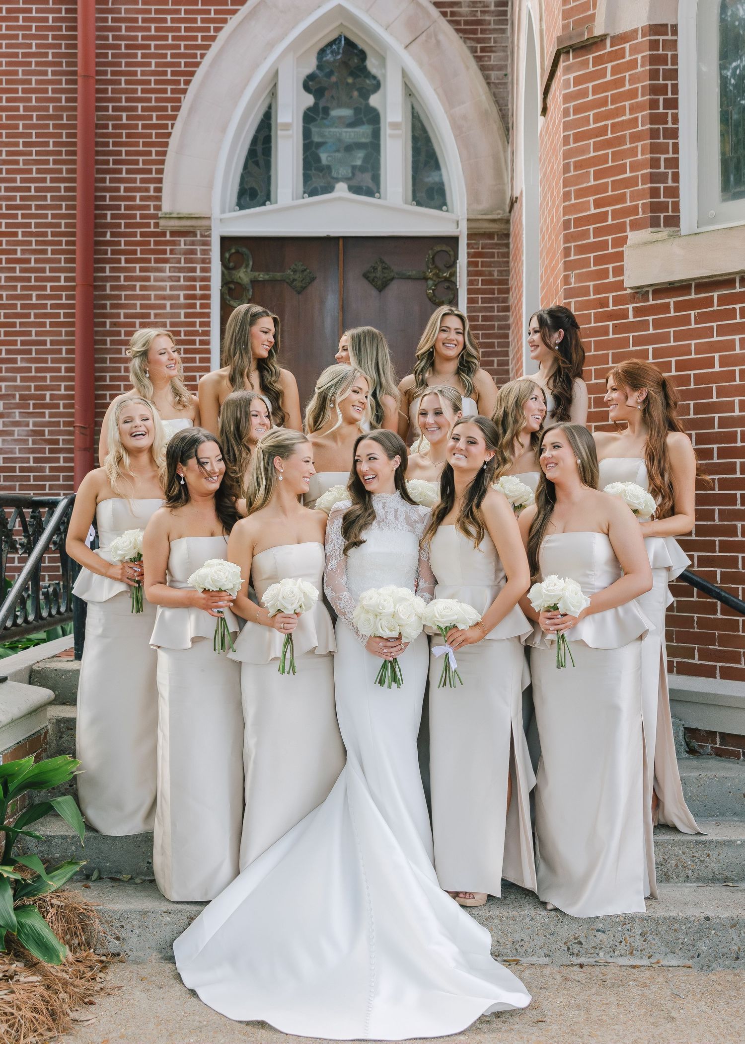 Bridesmaids in matching beige dresses gather around a bride in a white gown outside a brick church.
