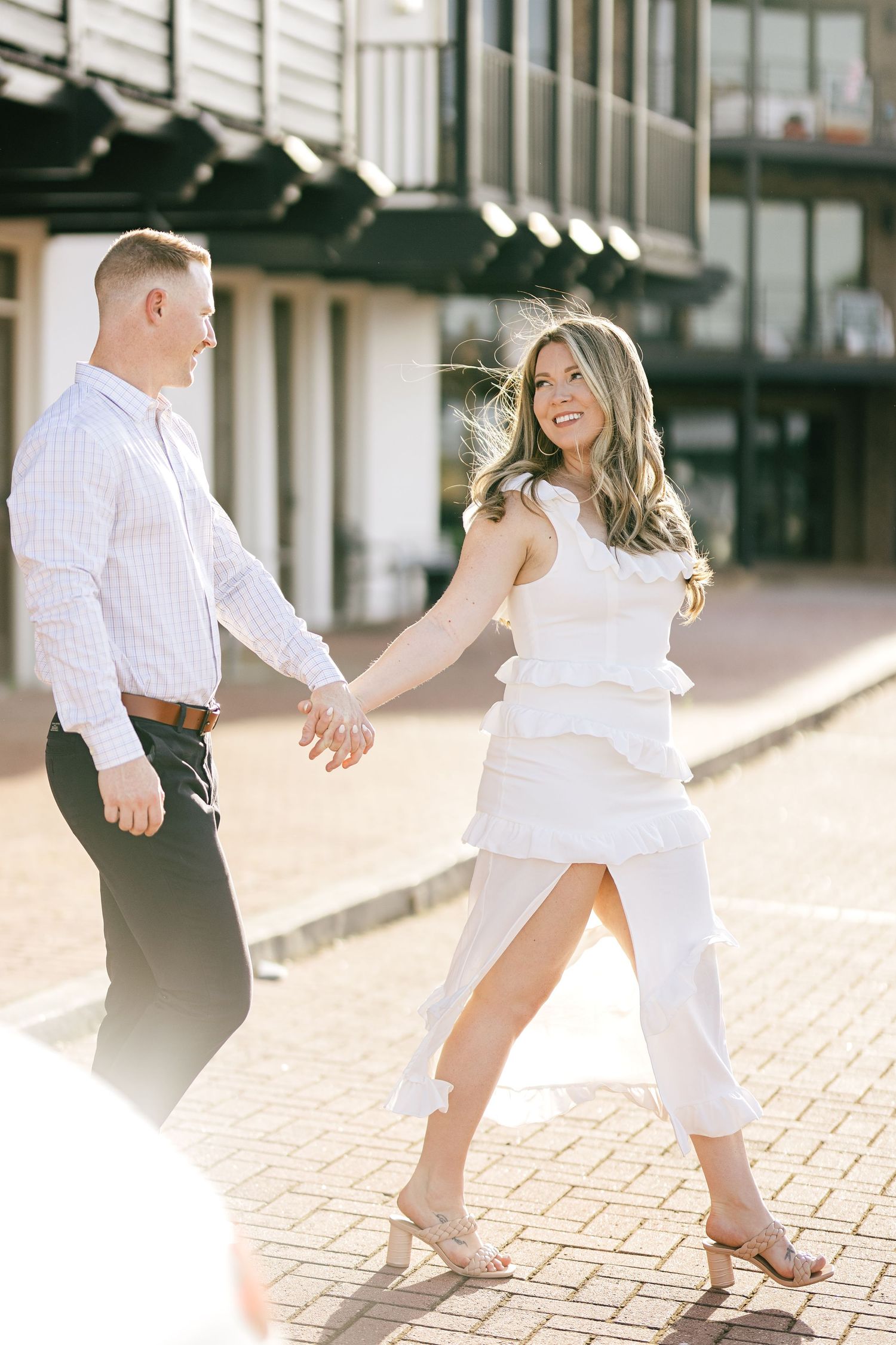 Playful couple in white outfits dancing together on brick pavement during a photo session.