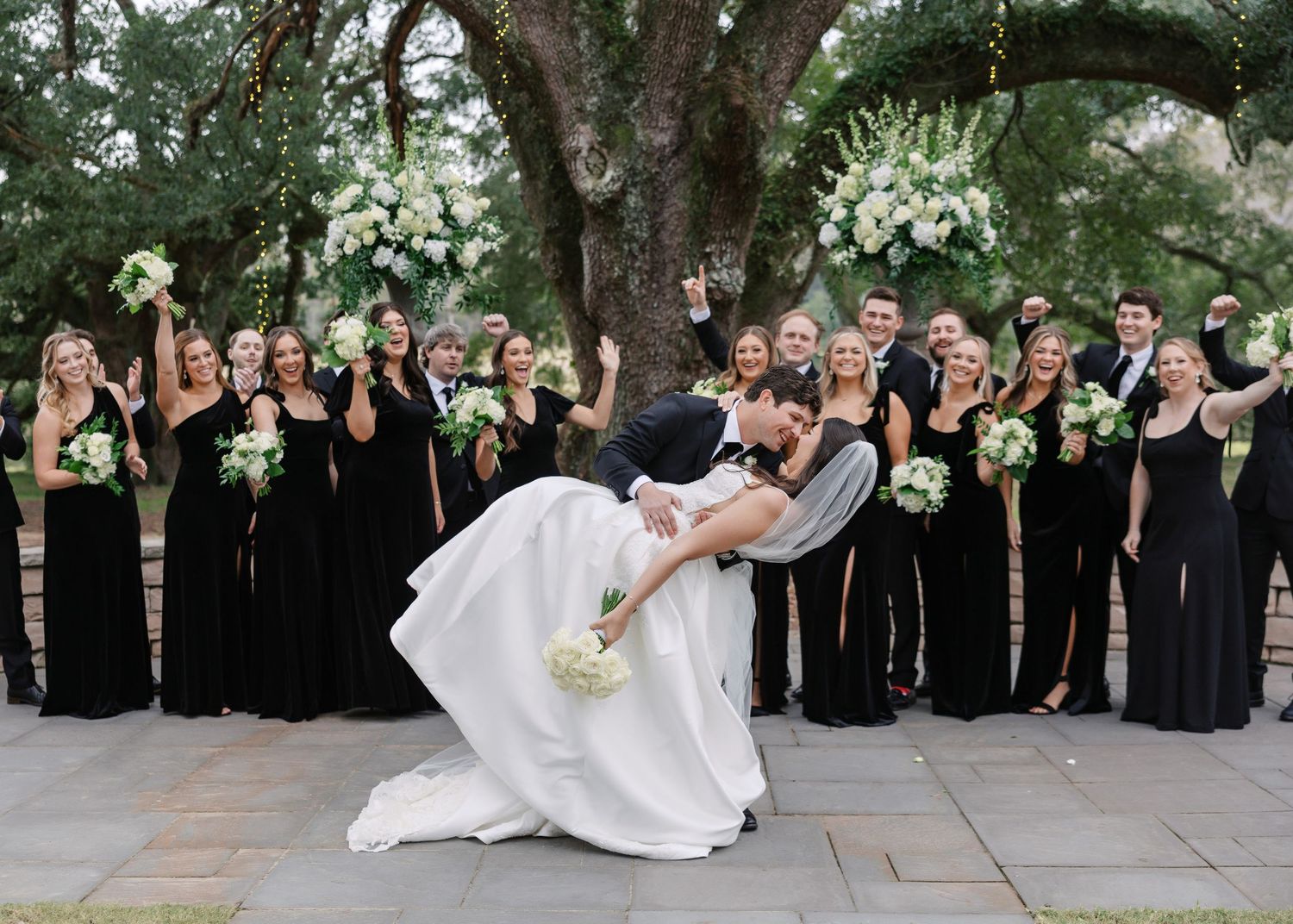 Wedding party in black attire celebrating with the couple on stone steps.