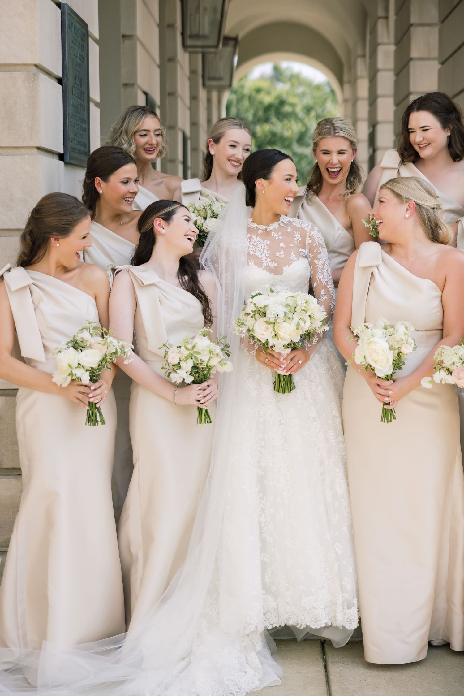 Bridesmaids in champagne dresses gather around bride in lace gown under arched walkway.