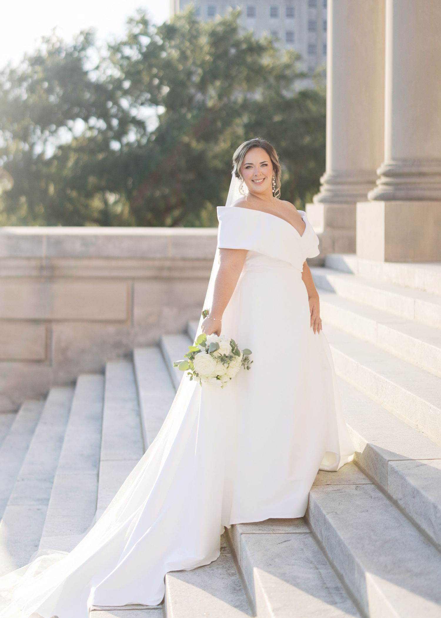 Elegant off-shoulder wedding gown photographed on grand stone steps in warm sunlight.