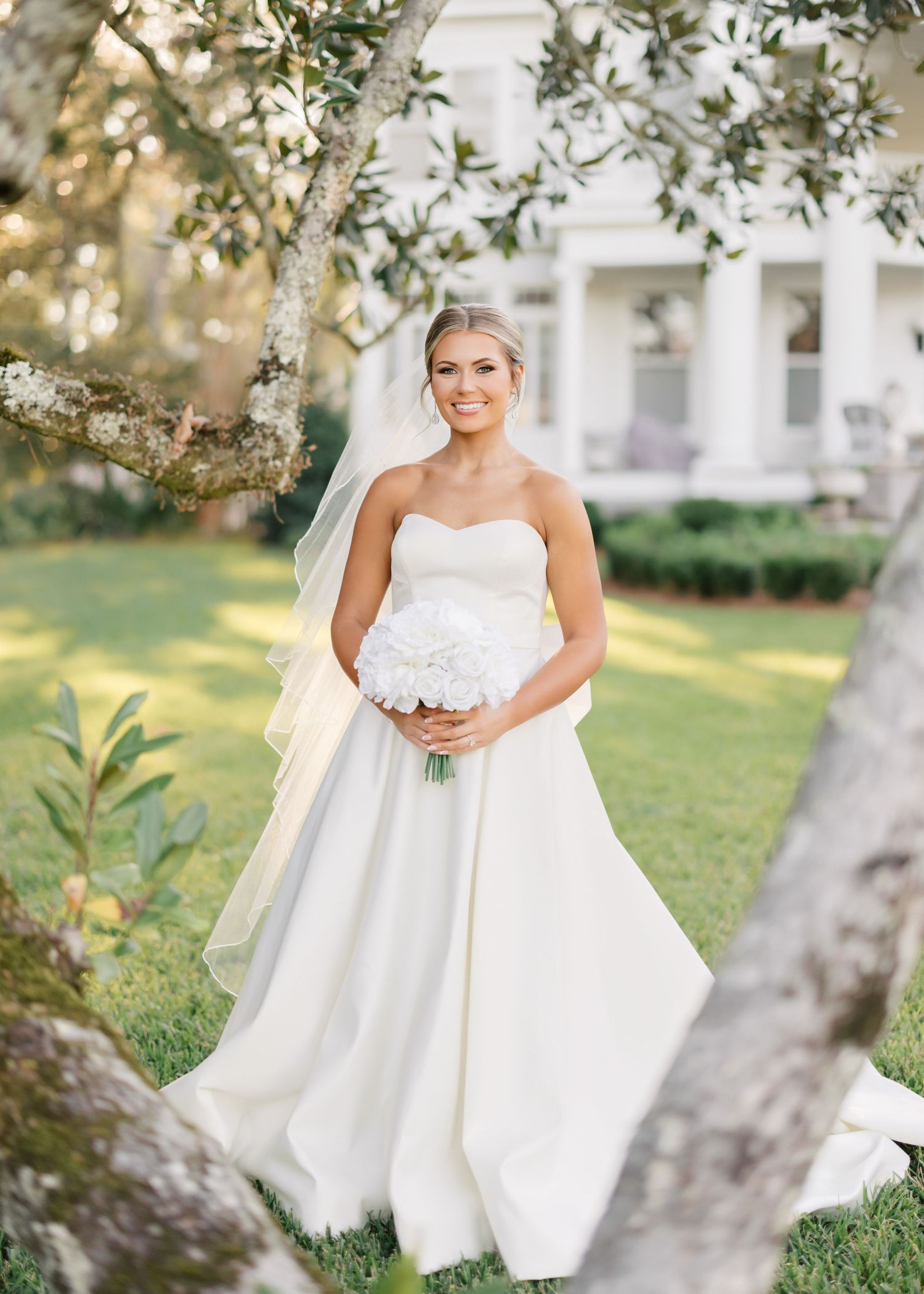 A bride in a flowing white wedding gown holds a bouquet while standing on a sunlit lawn near trees.