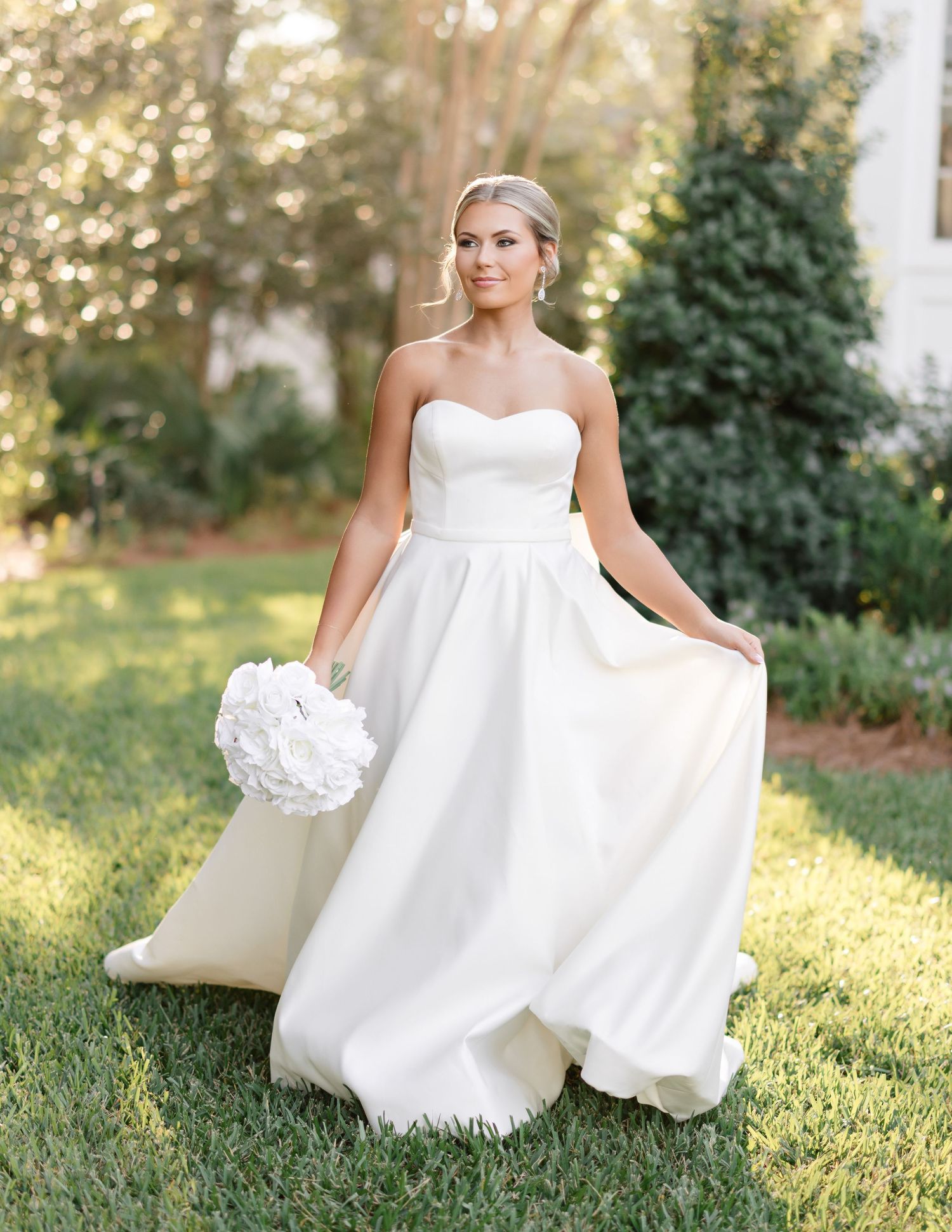 A bride in a flowing white gown sits gracefully on lush green grass during a sunny outdoor photoshoot.
