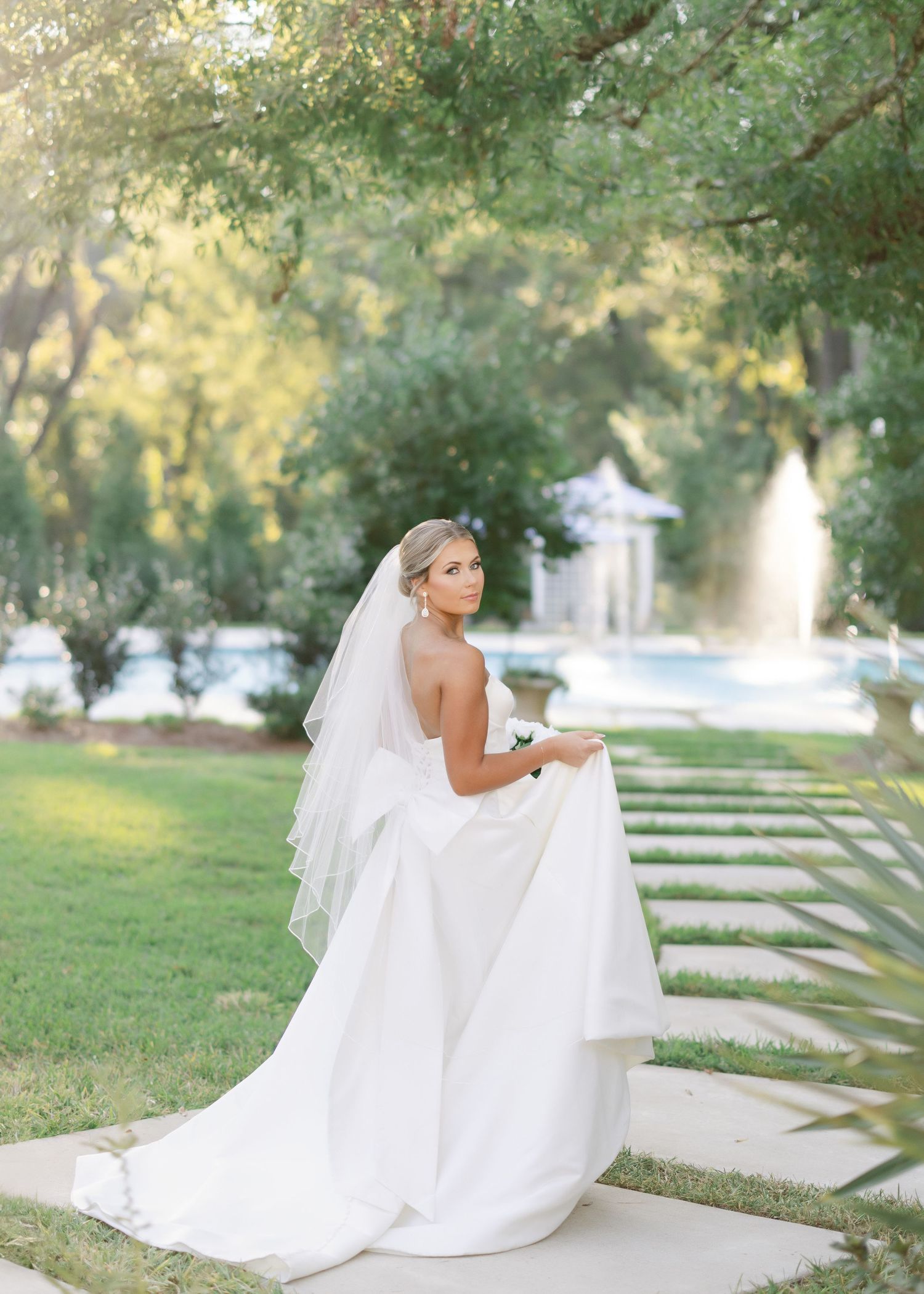 A bride in a flowing white wedding dress and veil poses gracefully in an outdoor garden setting at sunset.