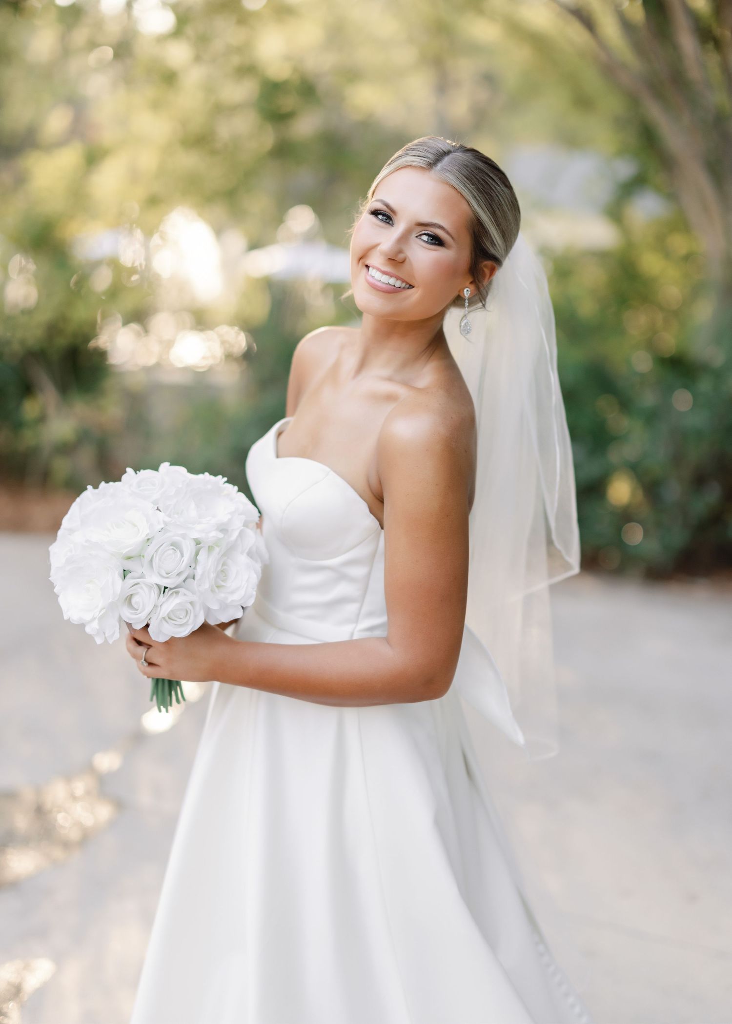 A bride in a white strapless gown holds a bouquet of white flowers while smiling outdoors in natural lighting.