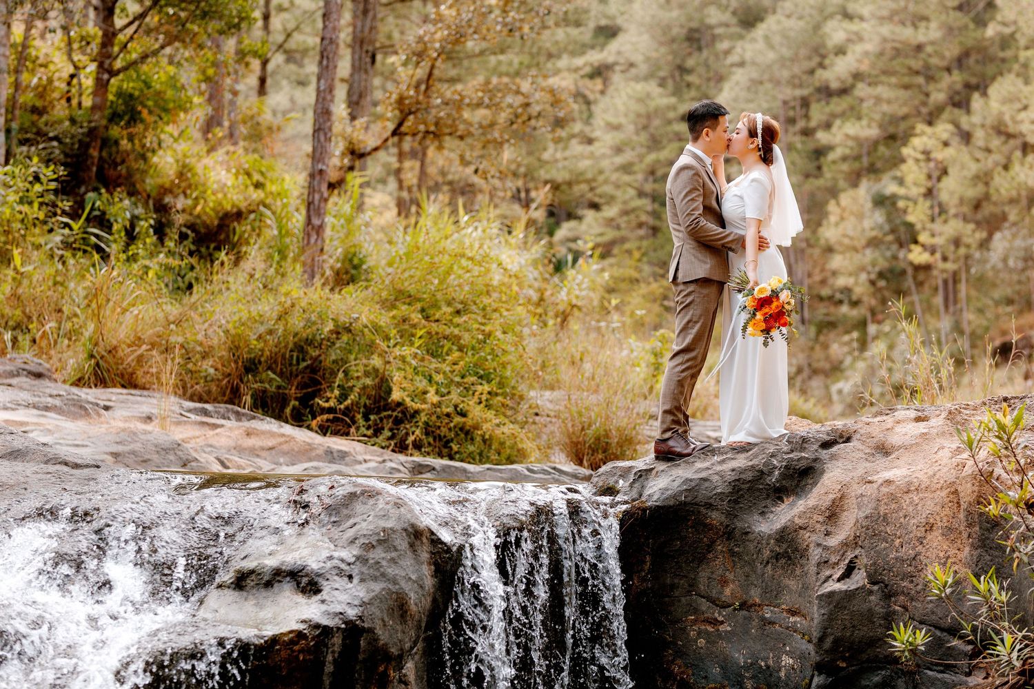 Bride and groom share a romantic kiss on a rocky waterfall edge surrounded by lush forest trees.