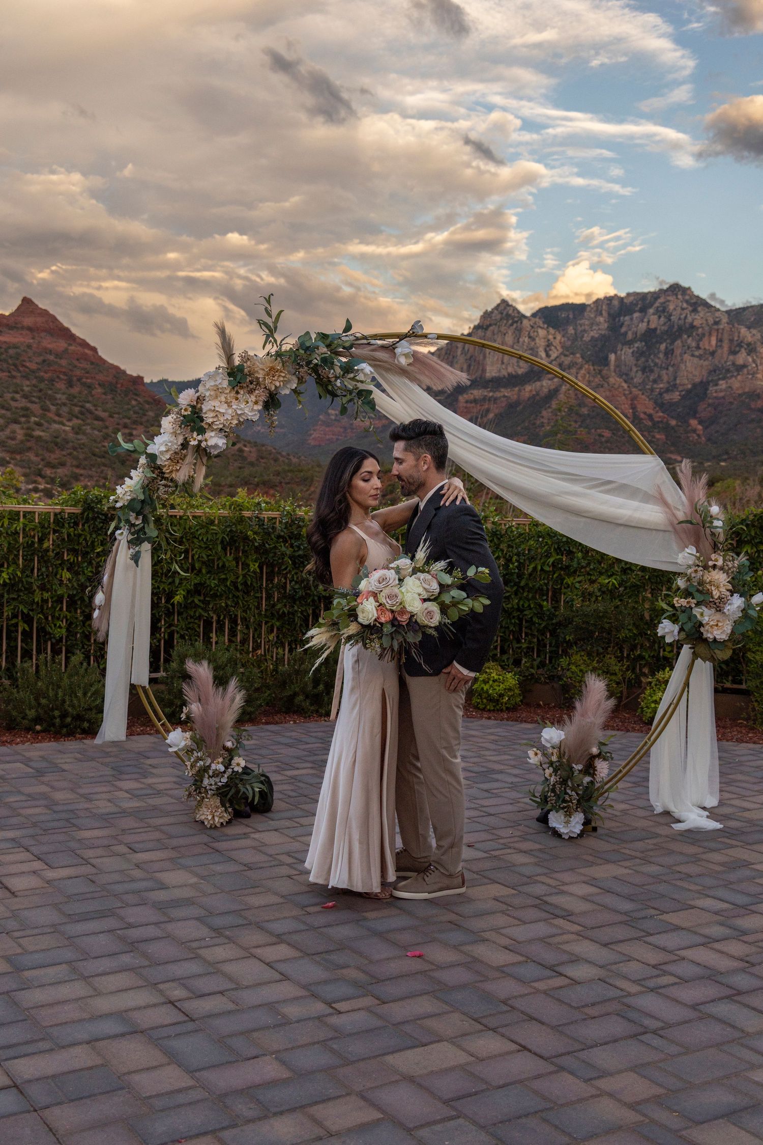 Outdoor wedding ceremony setup with white draped arch against dramatic mountain backdrop at sunset.