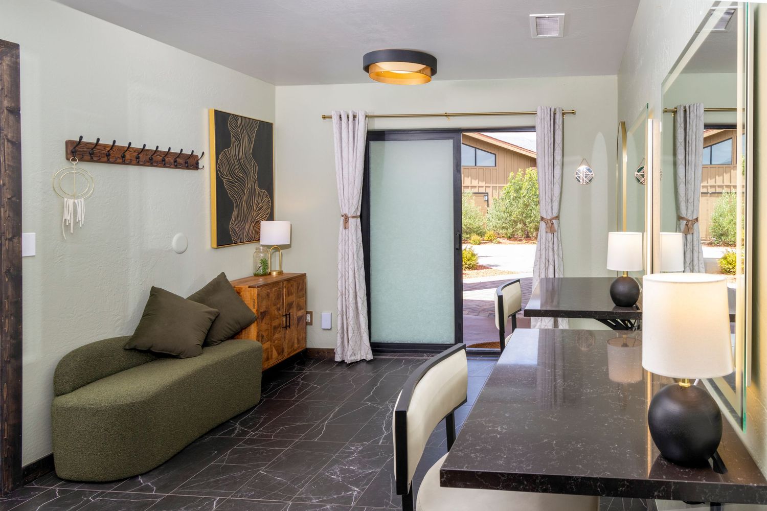 Modern hotel suite with green sofa, dark tile floors, frosted glass door, and elegant desk area with lamp.