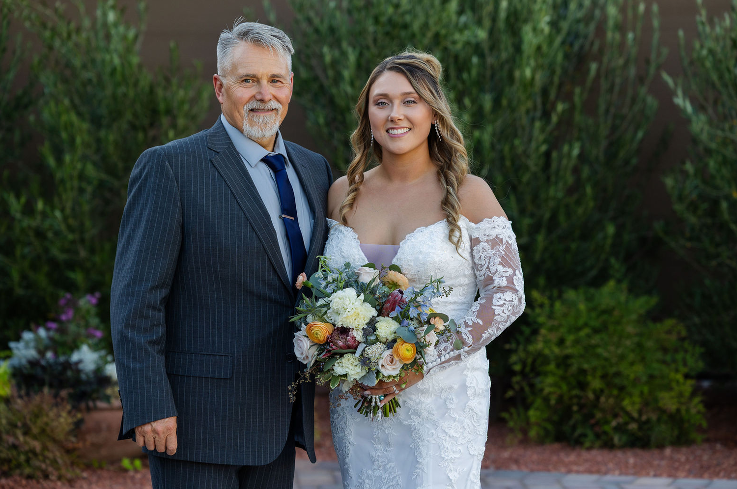 Bride in lace gown holding colorful bouquet poses with older man in navy suit at outdoor wedding ceremony.