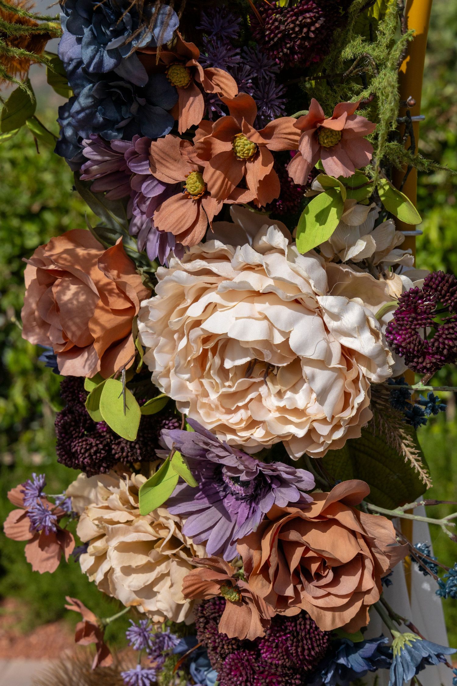 Colorful artificial flower arrangement featuring cream peonies, terracotta roses, purple daisies, and dark burgundy blooms outdoors.