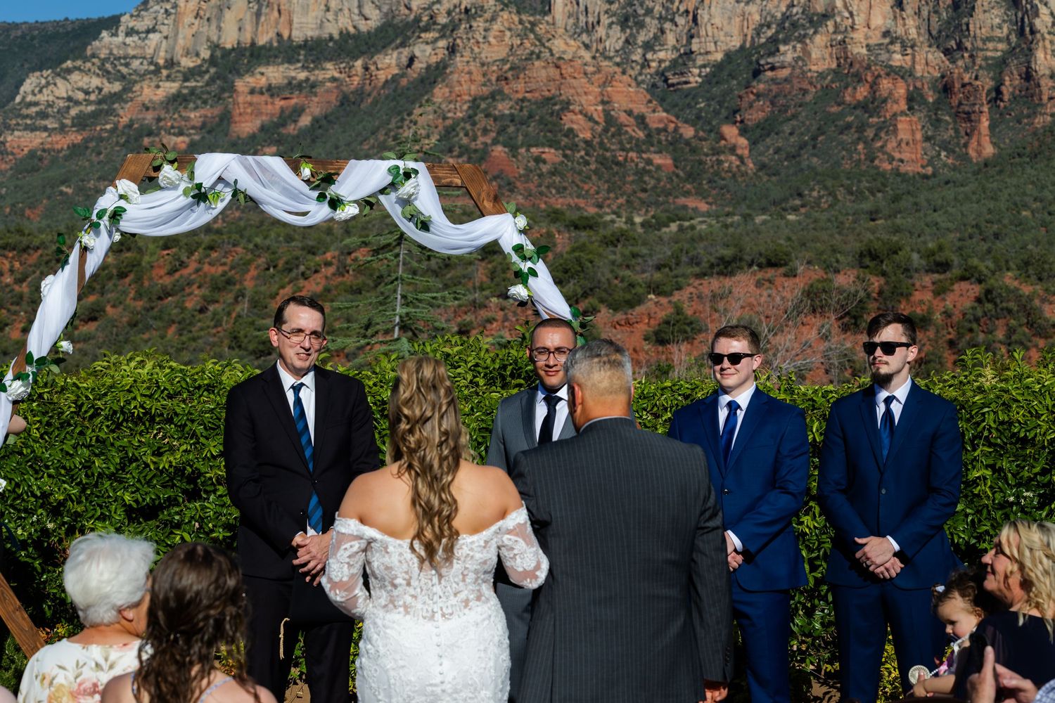 Outdoor wedding ceremony in Sedona Arizona with red rock mountains backdrop, bride groom and groomsmen at floral arch.