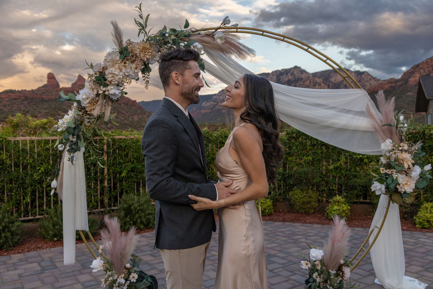 A couple embraces under a floral wedding arch at sunset with red rock mountains in the background at Sedona.