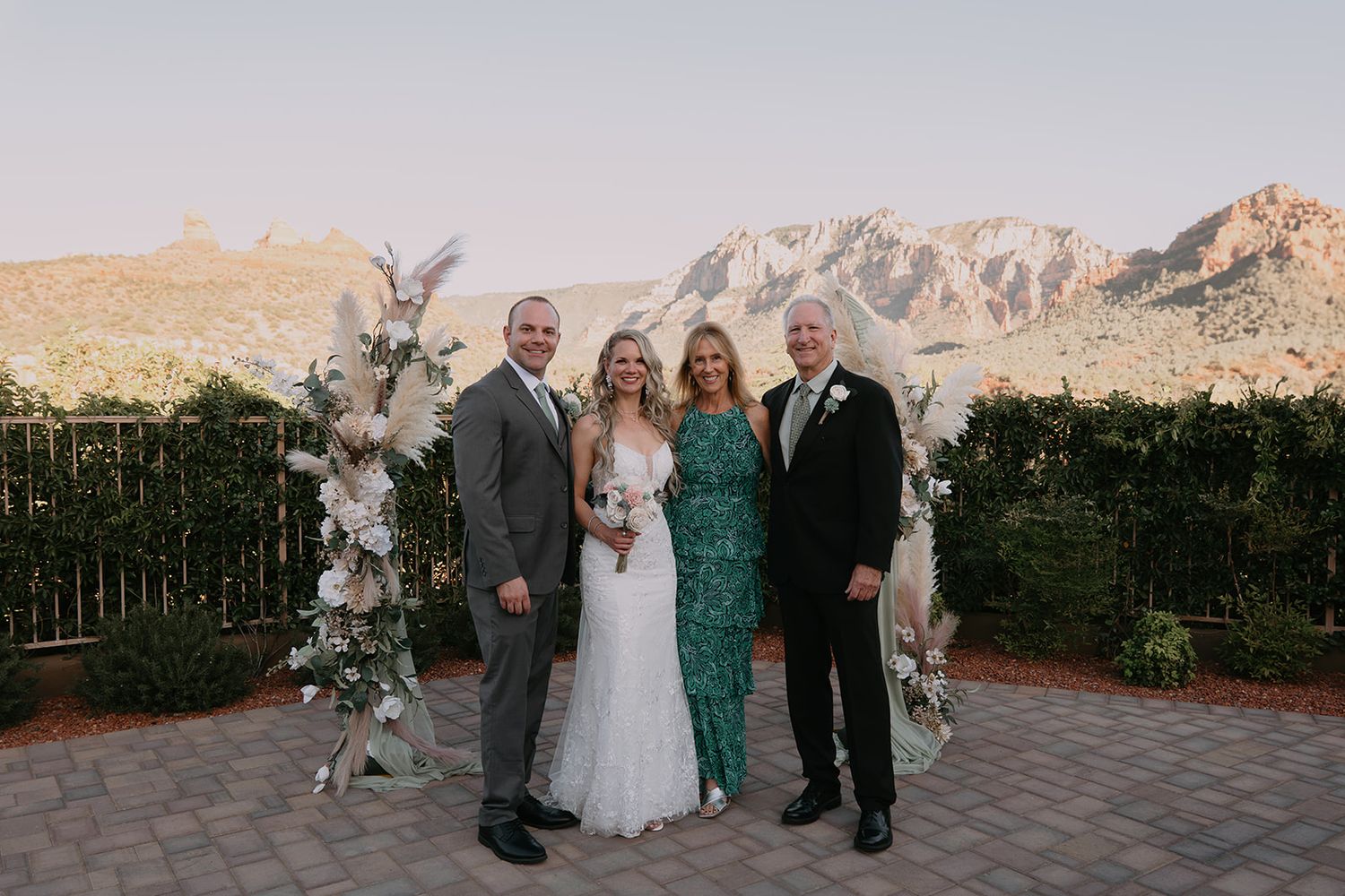 Bride and groom with family posing outdoors at desert wedding venue with mountain backdrop and floral arch.