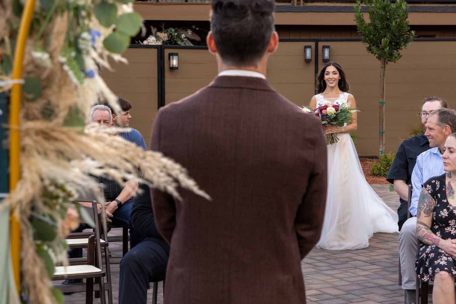 Groom in brown suit watches bride in white gown walk down outdoor aisle holding bouquet at wedding ceremony.