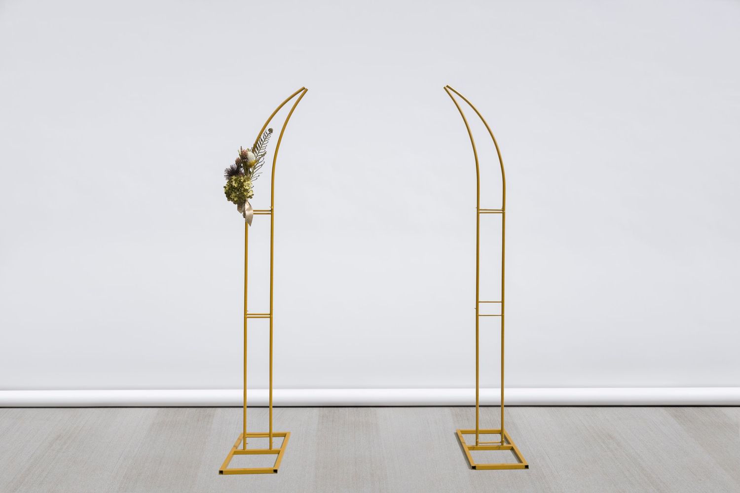 Gold curved metal arch frame stands against white wall with floral arrangement on top.