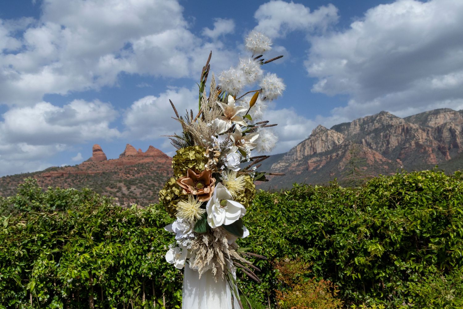 Floral arrangement with white blooms against red rock formations and lush green vineyard under cloudy blue sky.